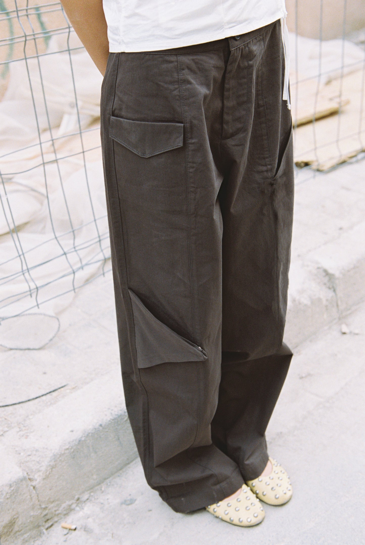 A person stands on a sidewalk near a wire fence and construction materials, wearing the Pocket Pant - Wood in a loose fit with dark cargo pockets and light-colored studded flats. Their upper body is not visible in the image.