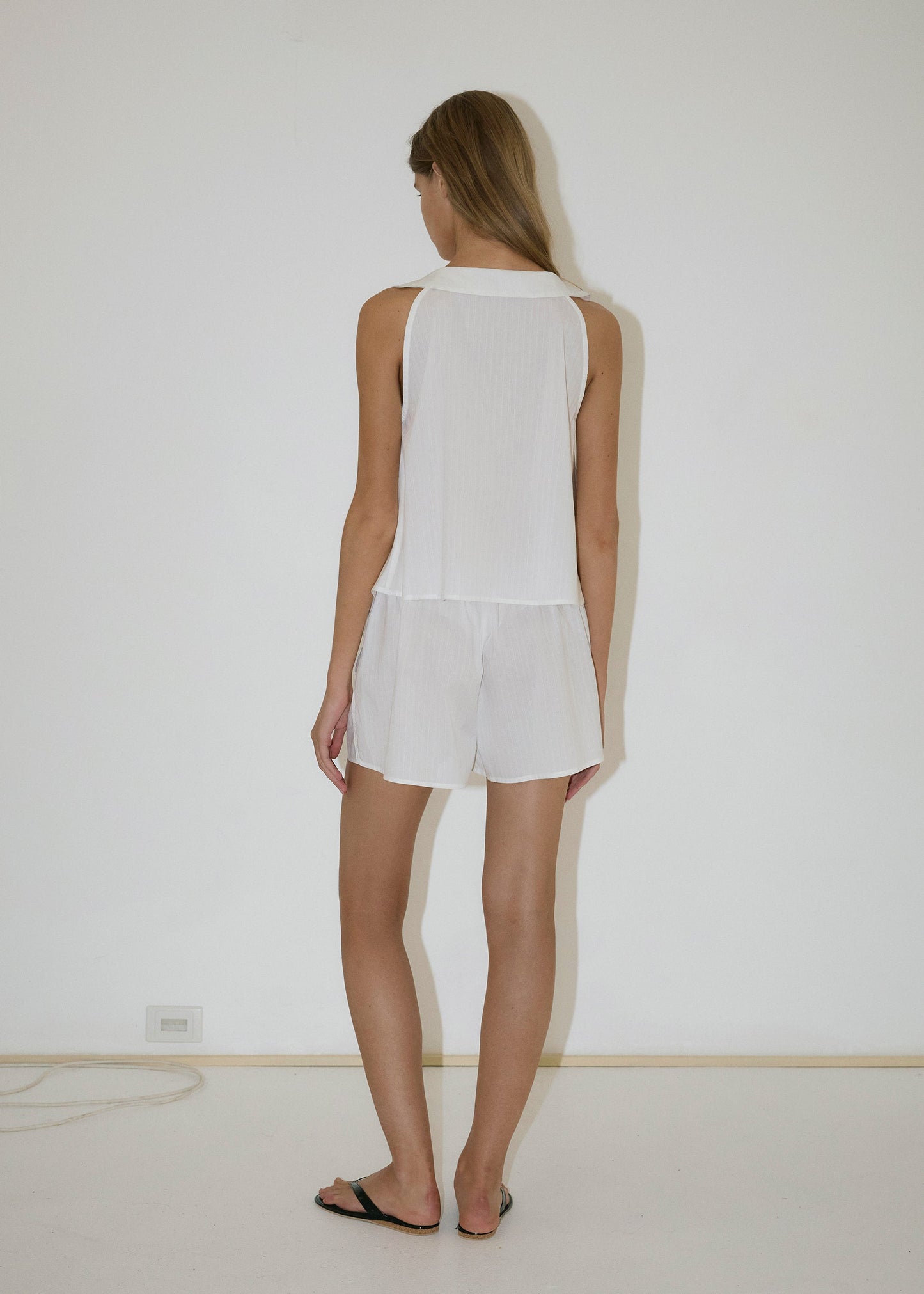 A person with long hair stands by a plain white wall, wearing the Round Collar Set - White Texture with an elastic waist and black sandals in a minimal space with a bare floor and visible outlet.