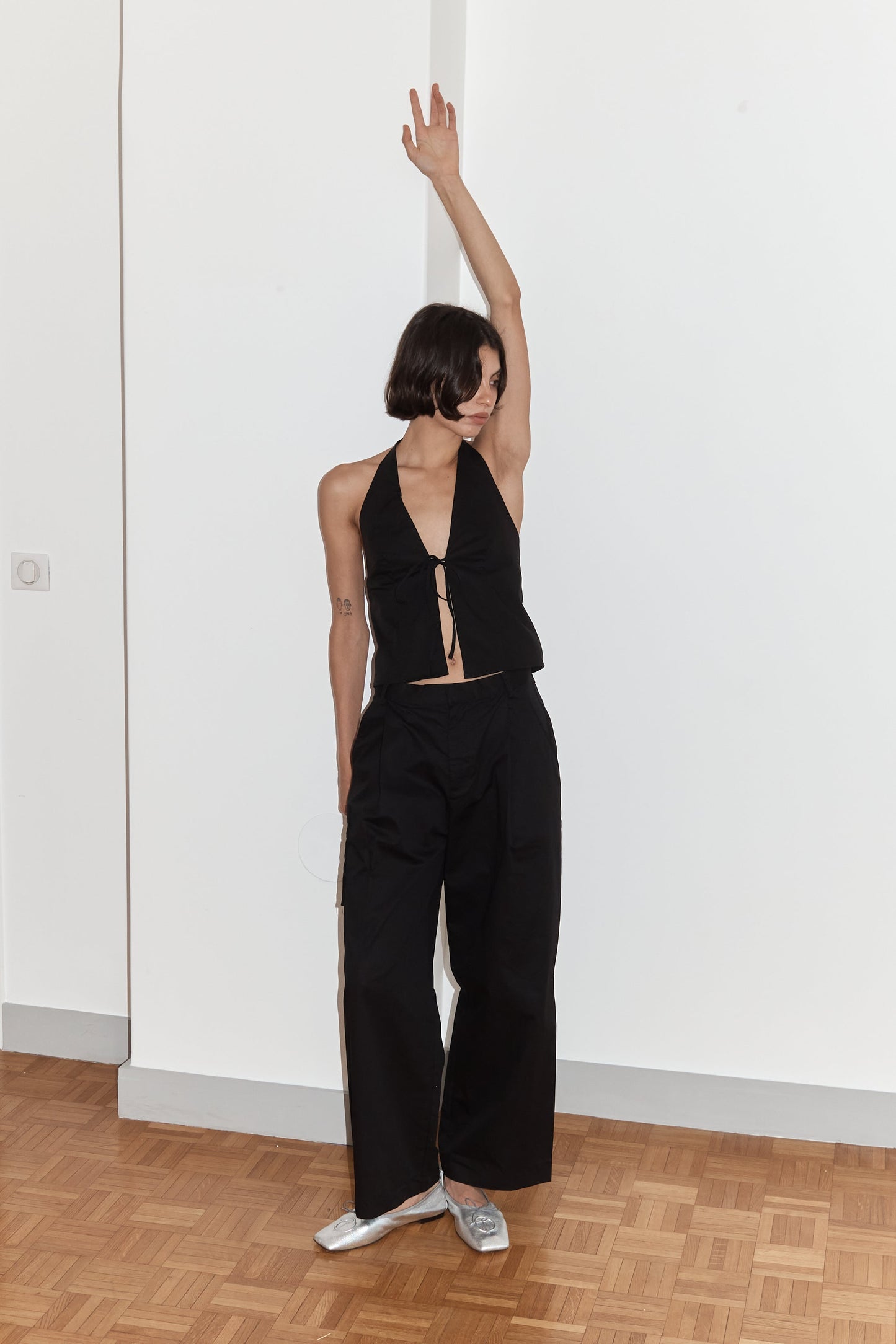 A woman with short dark hair stands on a wooden floor indoors, wearing the Open Halter Top - Black from Deiji Studios. She raises one arm overhead and looks up while her other arm hangs by her side.