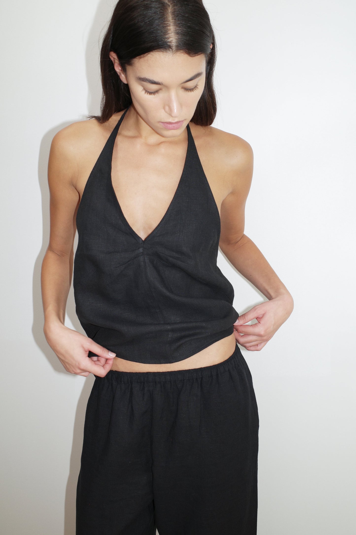 A woman with long dark hair wears the Linen Pinched Halter Top - Black and matching loose black pants, standing against a plain white background while looking down and adjusting her top.