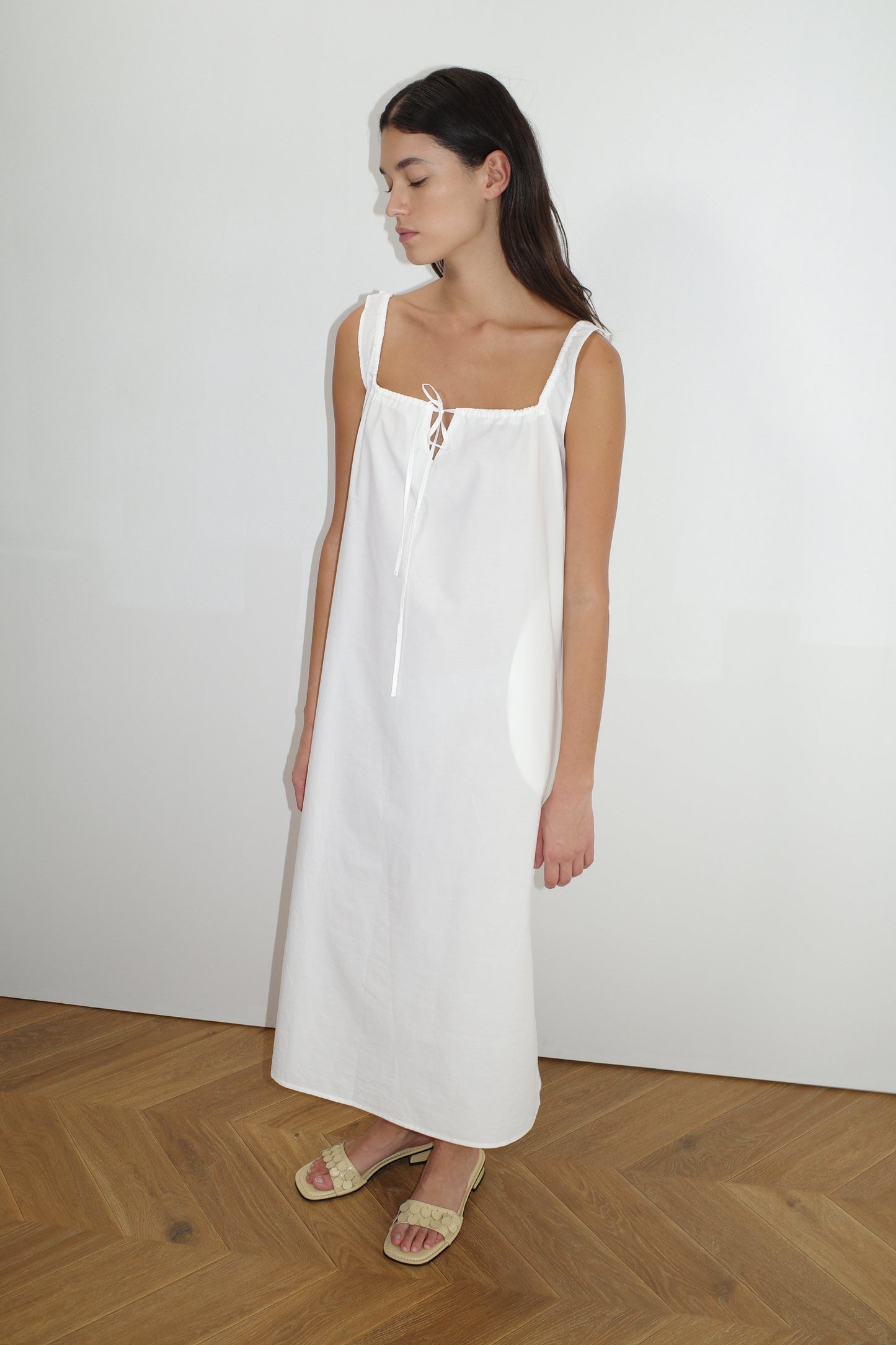 Model wearing The Paper Dress - White by Deiji Studios, featuring a square neckline, front tie, and wide straps, paired with beige woven slide sandals on herringbone wood flooring.