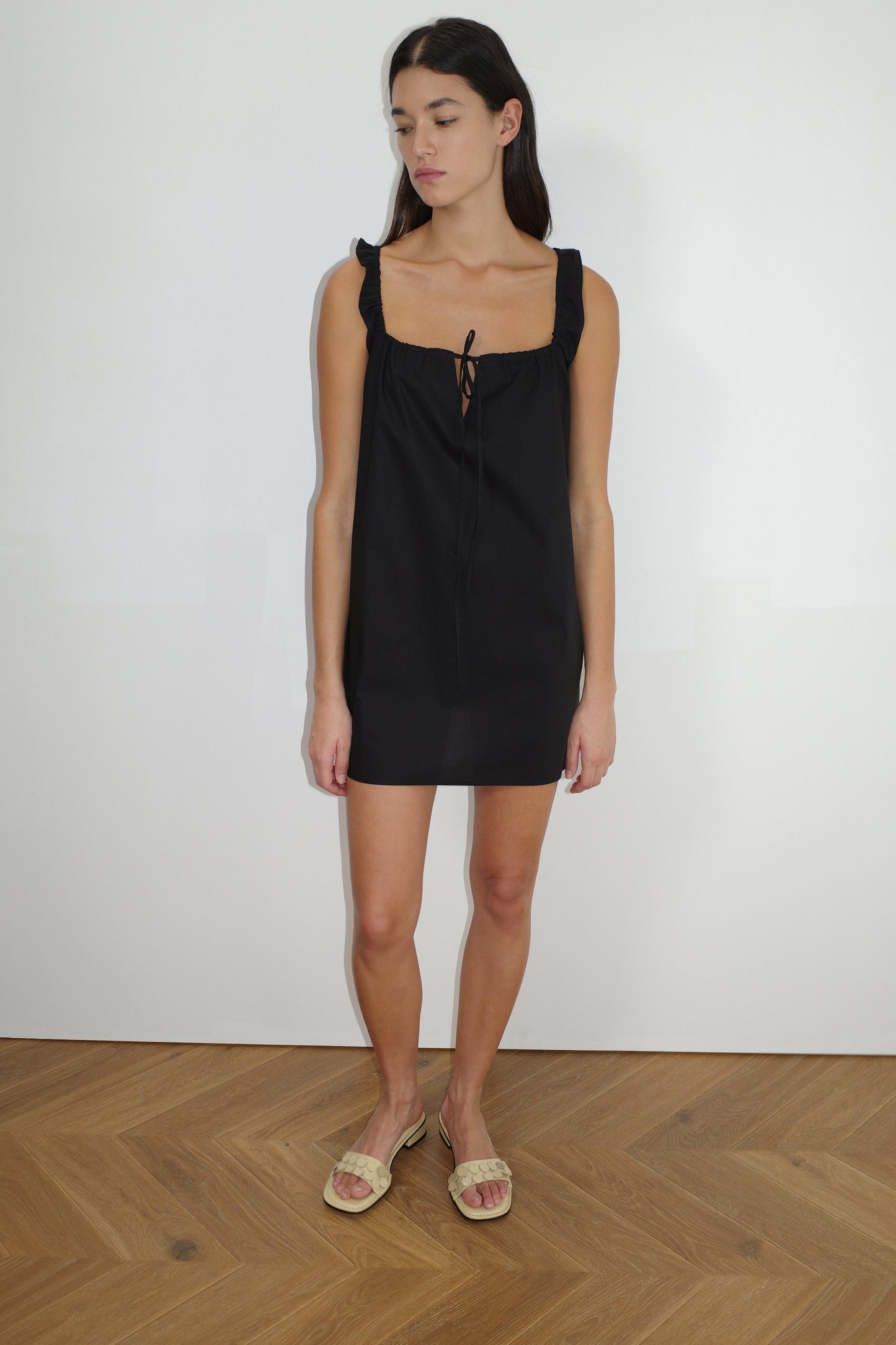 A woman with long dark hair stands indoors on a wooden floor, wearing The Bow Dress - Black by Deiji Studios, featuring ruffled straps and a tie neckline, paired with beige open-toe sandals. She is looking slightly downward.