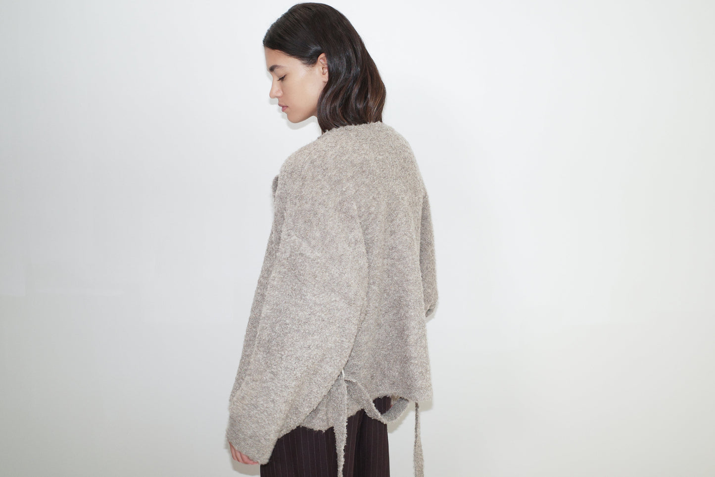 A person with shoulder-length dark hair wears the Pod Knit - Cloud Grey by Deiji Studios, a loose, textured sustainable sweater, paired with dark pants, standing sideways against a plain white background with their head slightly down.
