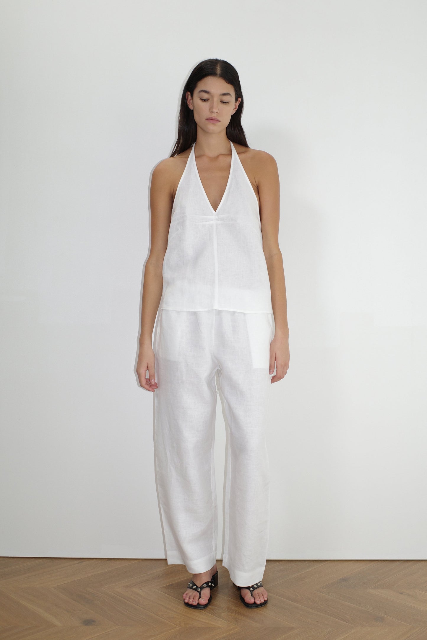 A woman with long dark hair stands indoors against a white wall, facing forward in a sleeveless white halter top, Linen Ease Trouser - White by Deiji Studios, and black sandals, wearing a neutral expression.