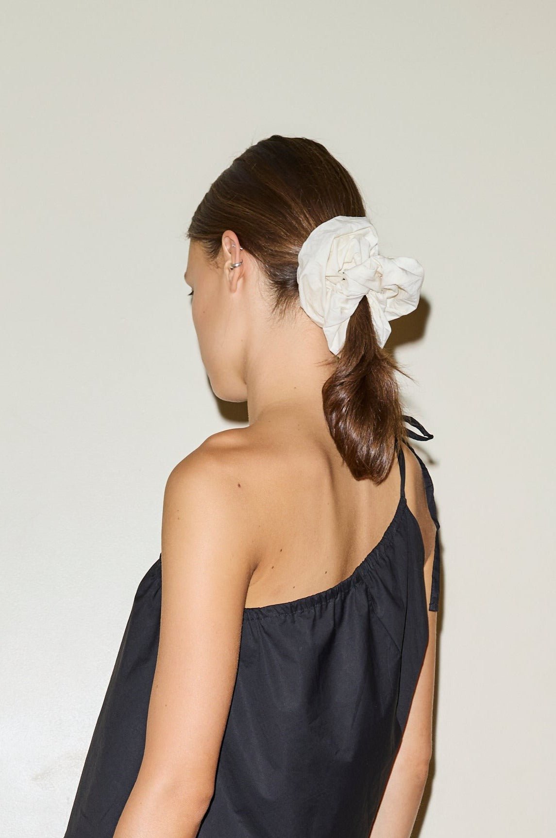 A woman with brown hair in a low ponytail wears a large white Scrunchie, paired with a black one-shoulder top, standing against a plain light wall and facing away from the camera.