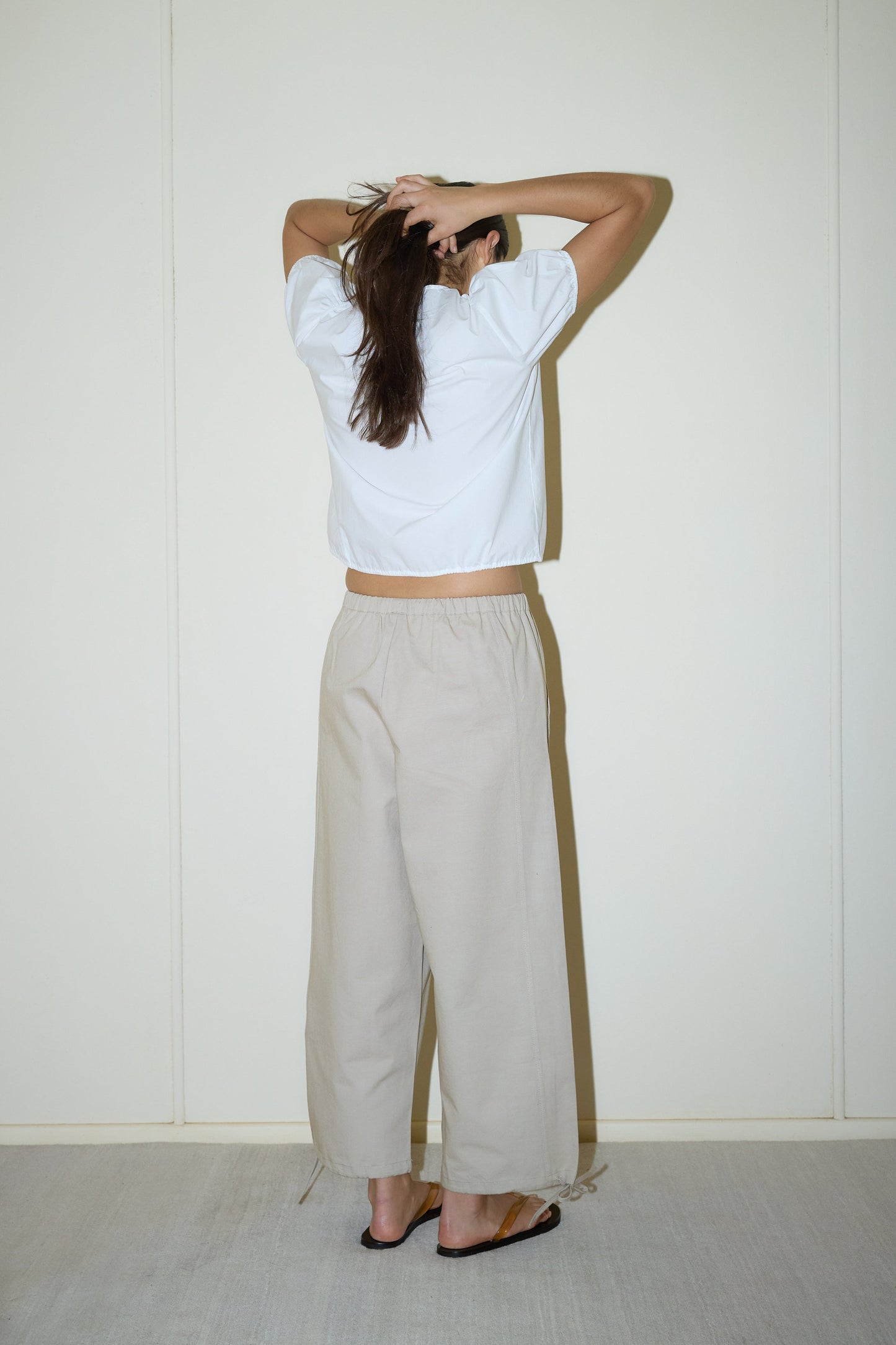 A person stands with their back to the camera facing a white wall, wearing a Double Frill Top - White from Deiji Studios, beige wide-leg pants, and sandals, with their hands raised to the back of their head.