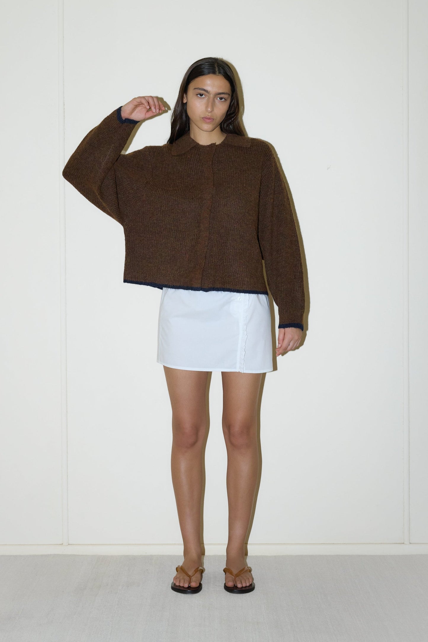 Model wears the One Button Knit - Tawny by Deiji Studios, an oversized brown sustainable knit cardigan with navy trim and collar, paired with a white mini skirt and brown leather flip-flops, standing against a cream wall.
