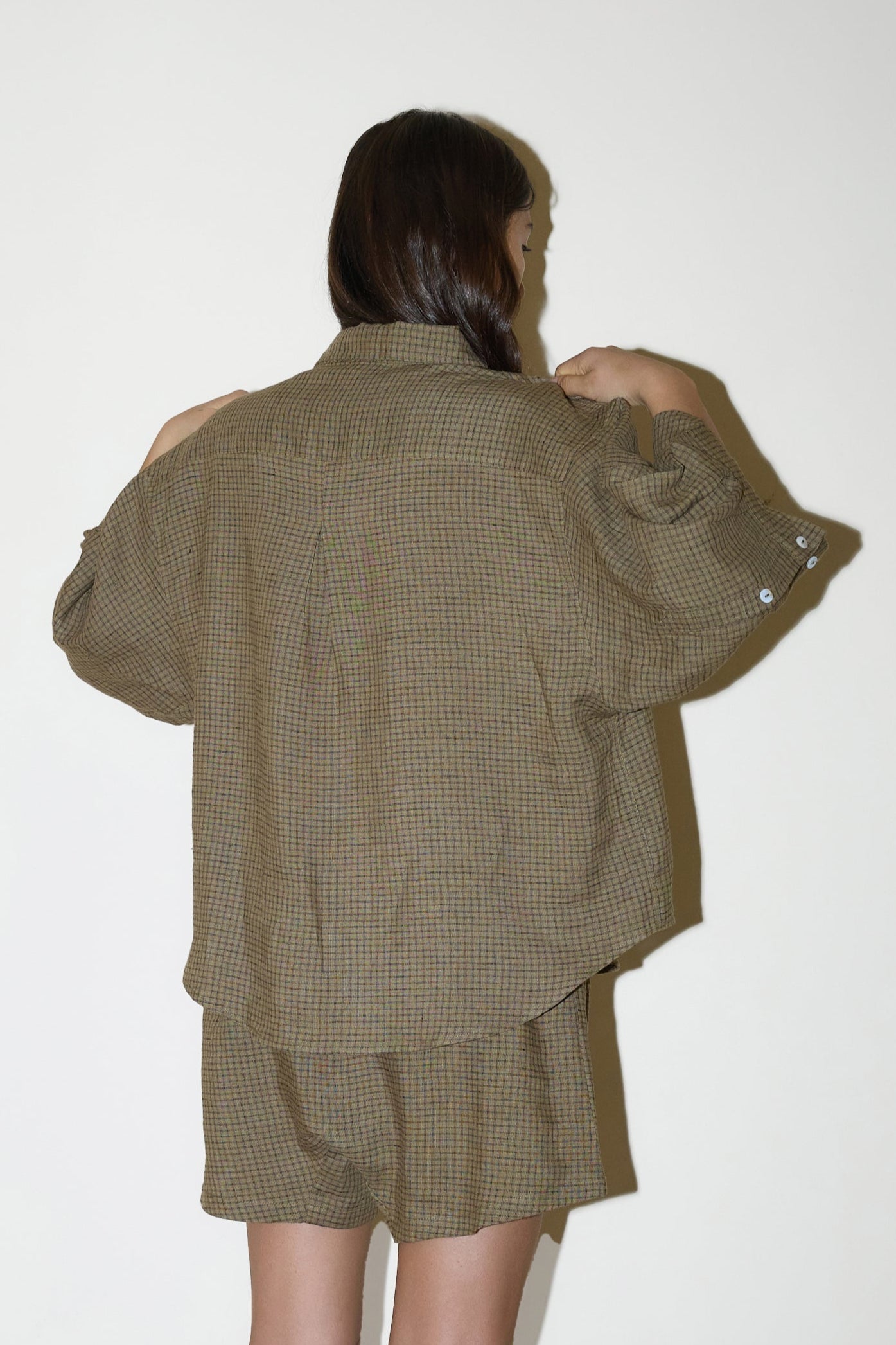 A person with long brown hair, seen from behind, wears the 03 Set - Pine Check: an oversized linen shirt and matching boxer-style shorts in brown plaid, standing against a plain light background.