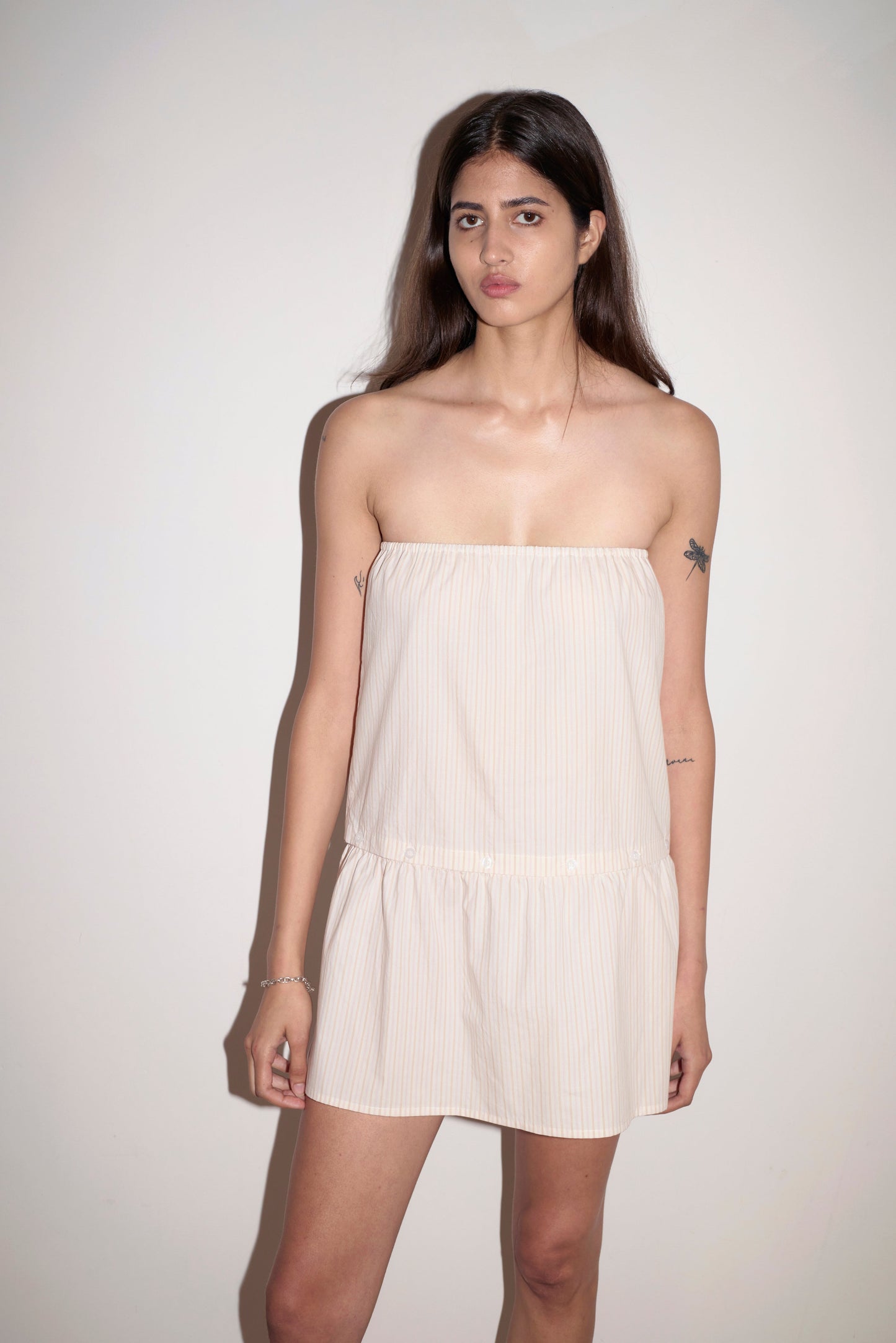 A woman with long dark hair stands against a plain light wall, wearing the Button Dress—a strapless, loose-fitting mini dress with subtle vertical stripes. She has a neutral expression and visible tattoos on her arms.