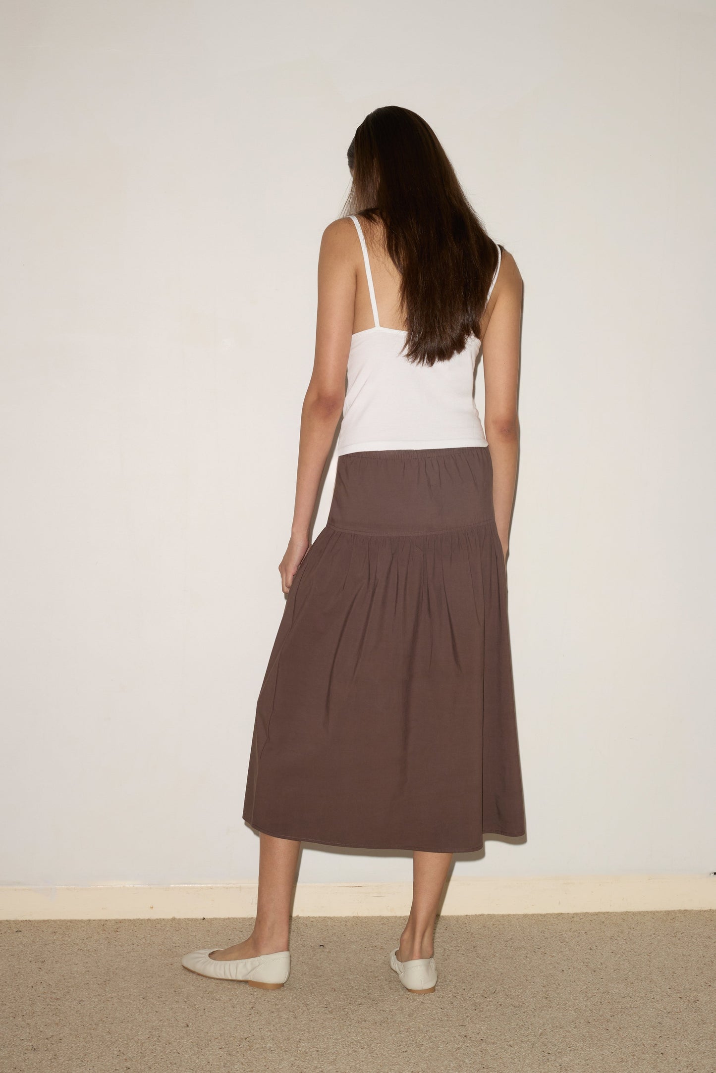 A woman with long brown hair stands facing away from the camera, wearing a Simple Tank - White by Deiji Studios, a brown midi skirt, and cream flat shoes against a plain light-colored wall and carpeted floor.