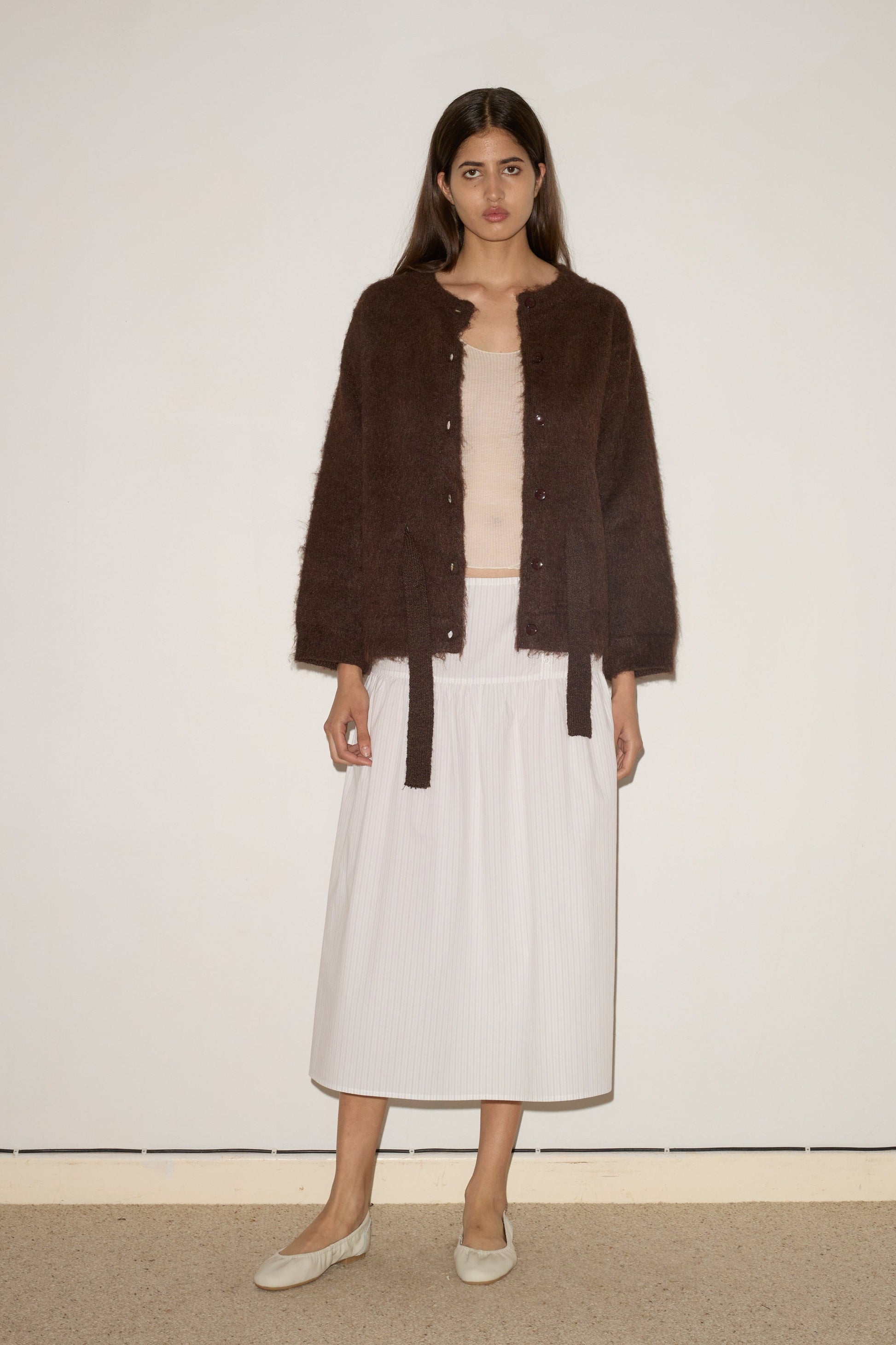 A woman with long dark hair stands against a plain wall wearing the 0 Belted Cardi - Brown over a light beige top, a white ankle-length skirt, and beige flat shoes.