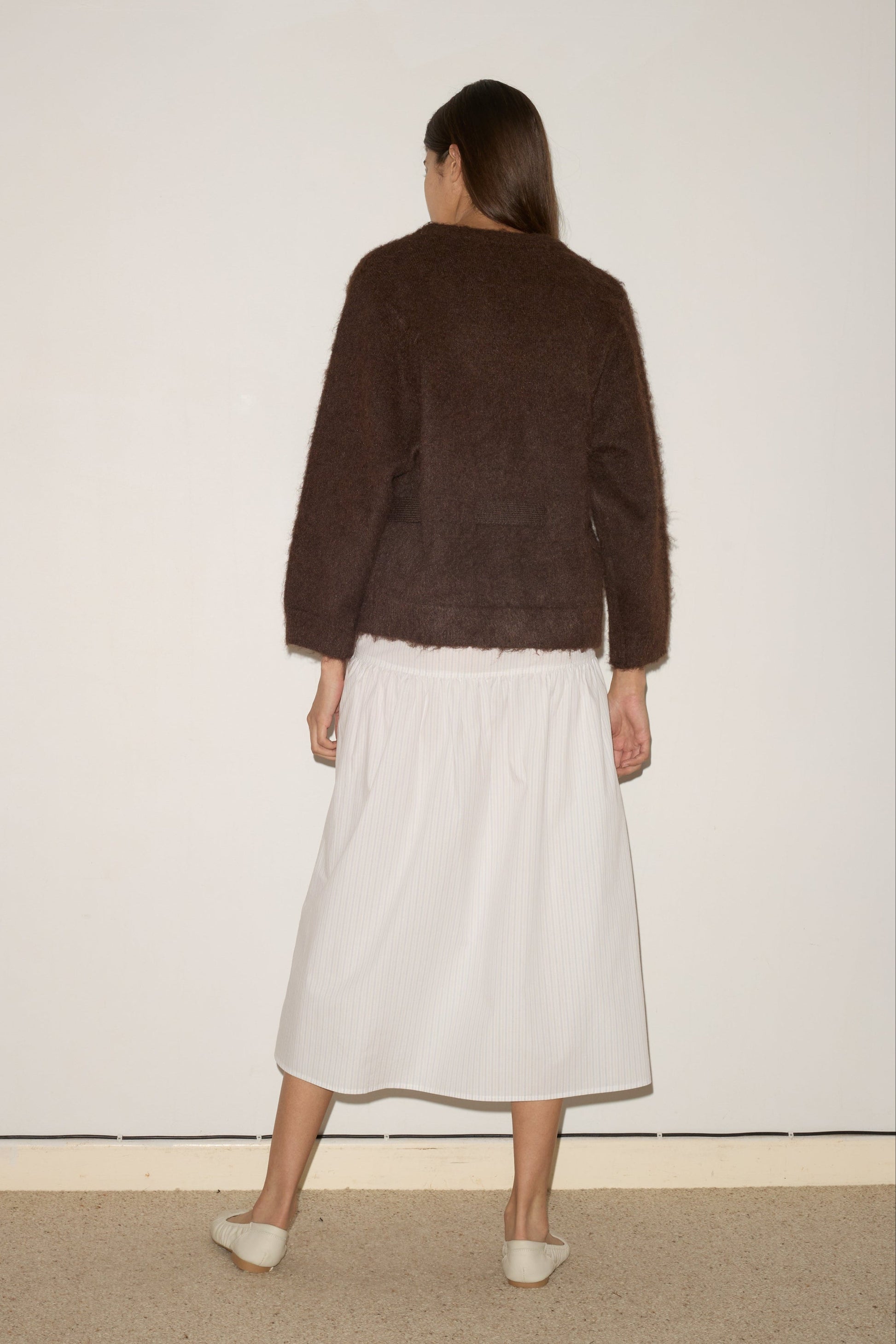 A woman with long hair stands facing a plain wall, seen from behind, wearing the 0 Belted Cardi - Brown over a white midi skirt and light-colored flats.