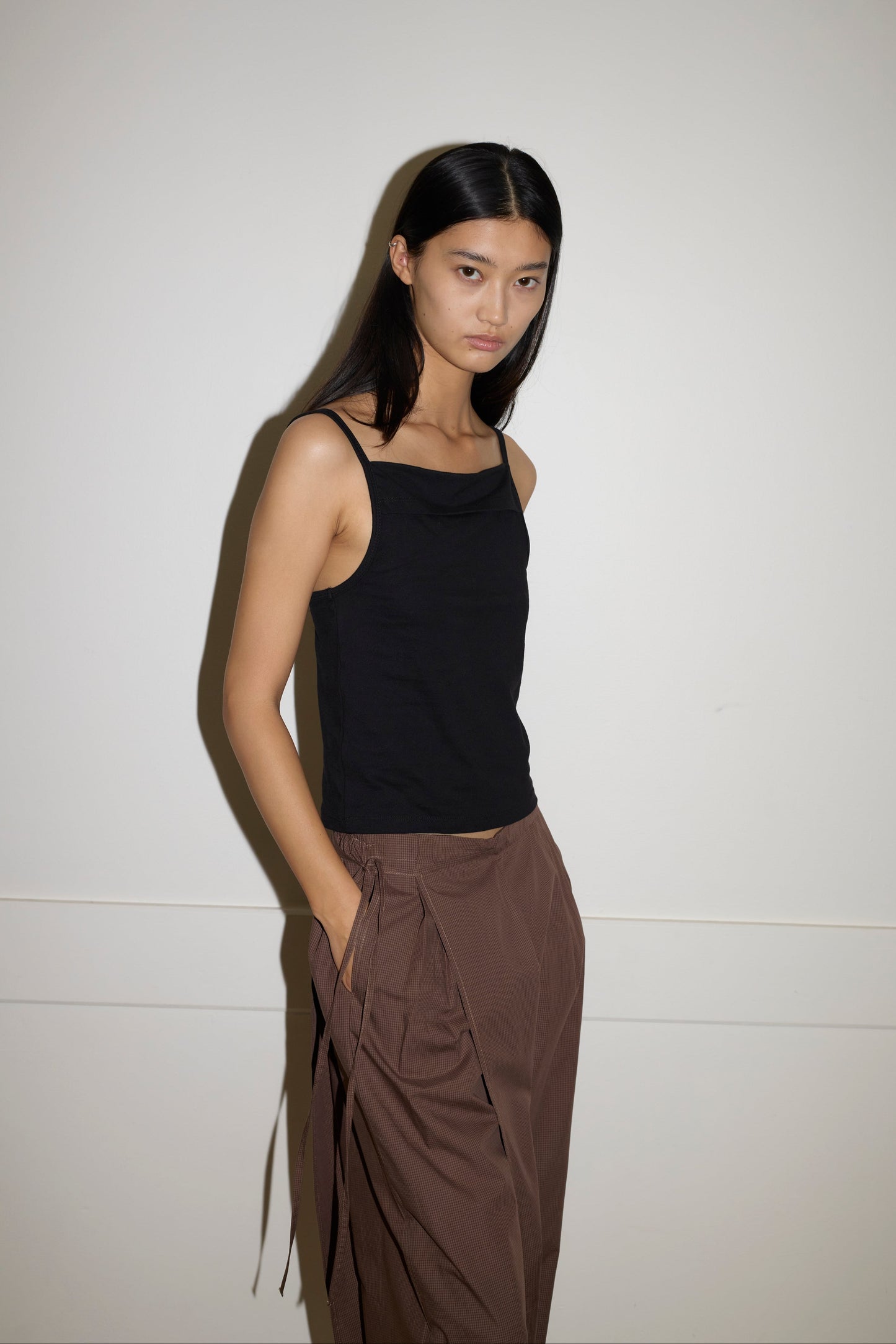 A woman with long black hair stands against a plain light wall, wearing a sleeveless black top and the Double Layer Pant - Carob Check, hands in her pockets, looking at the camera with a neutral expression.