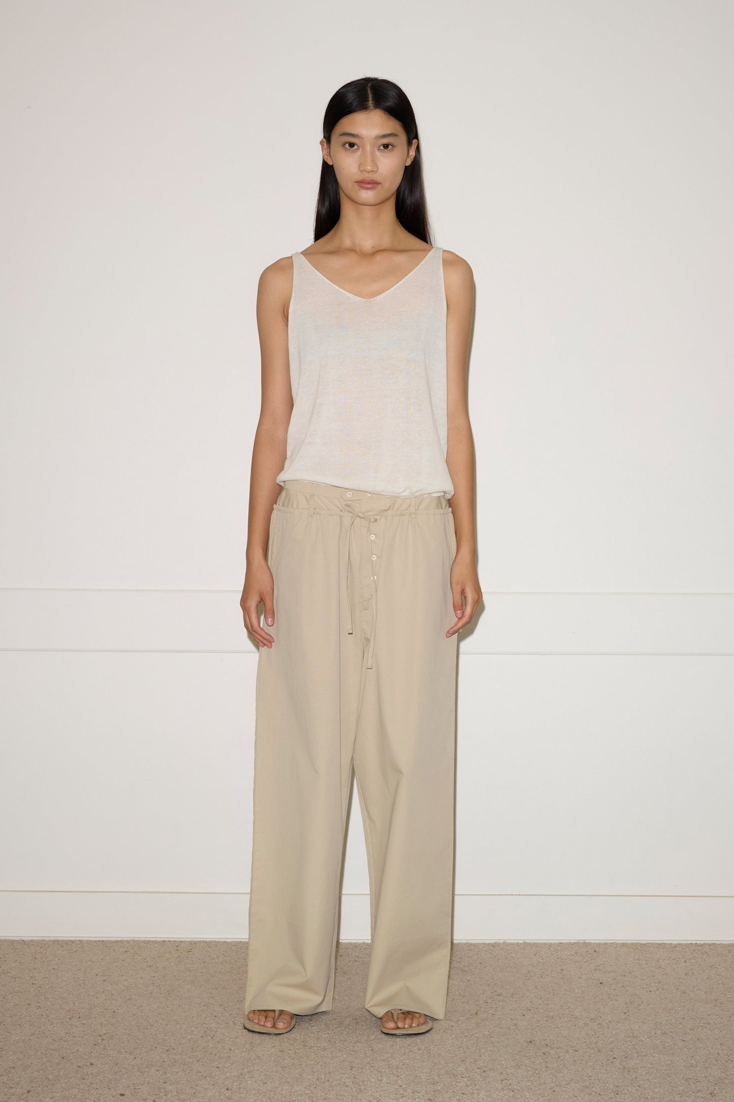 A person stands against a plain white wall, wearing Deiji Studios organic cotton sleeveless top and the Two Band Pant - Fawn with open-toed sandals. The expression is neutral and their relaxed pose features arms at their sides.