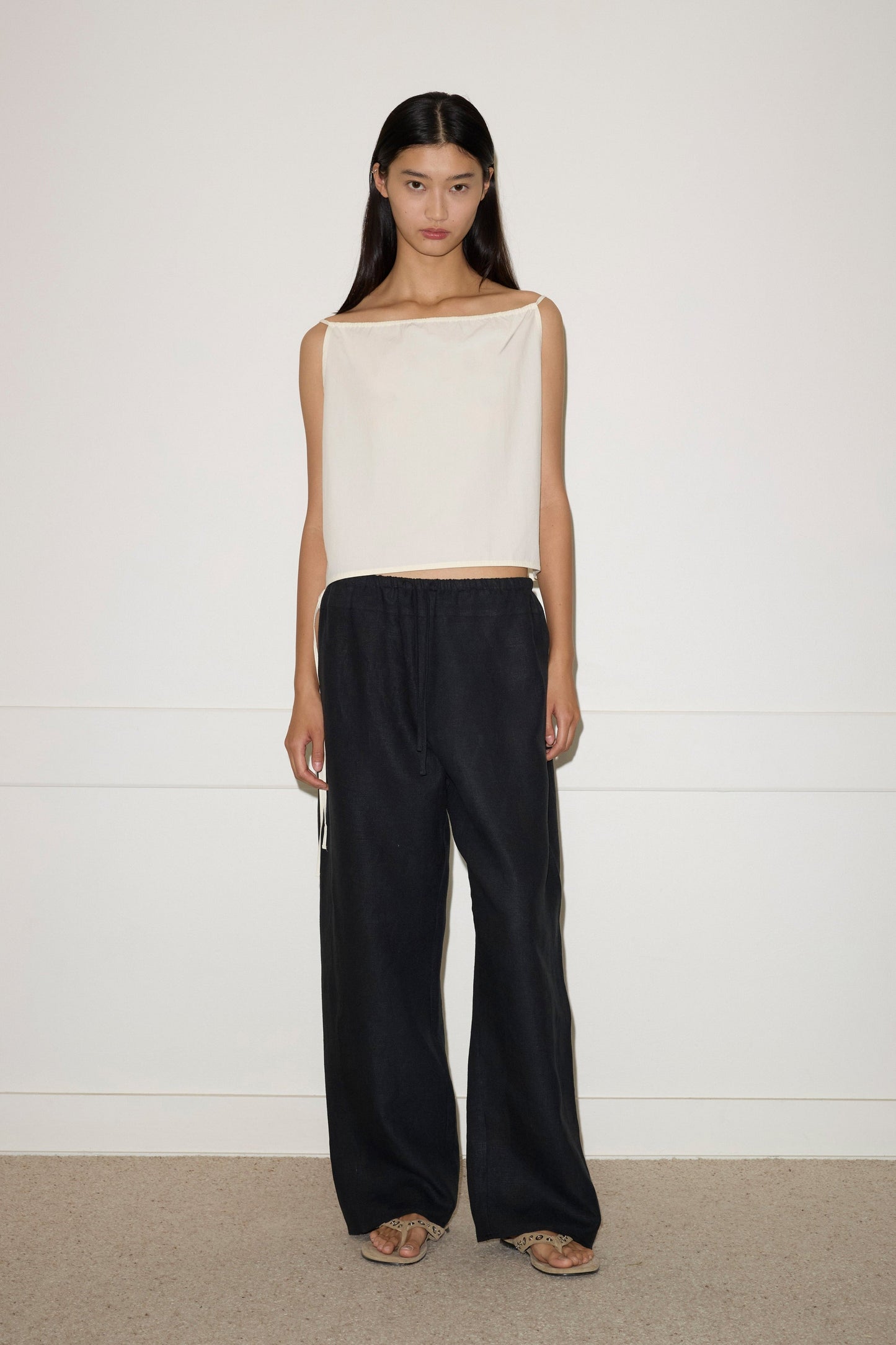 A woman with long dark hair stands by a white wall, wearing sandals and wide-leg black Double Waist Linen Pants with drawstrings, paired with a loose, sleeveless off-white top.
