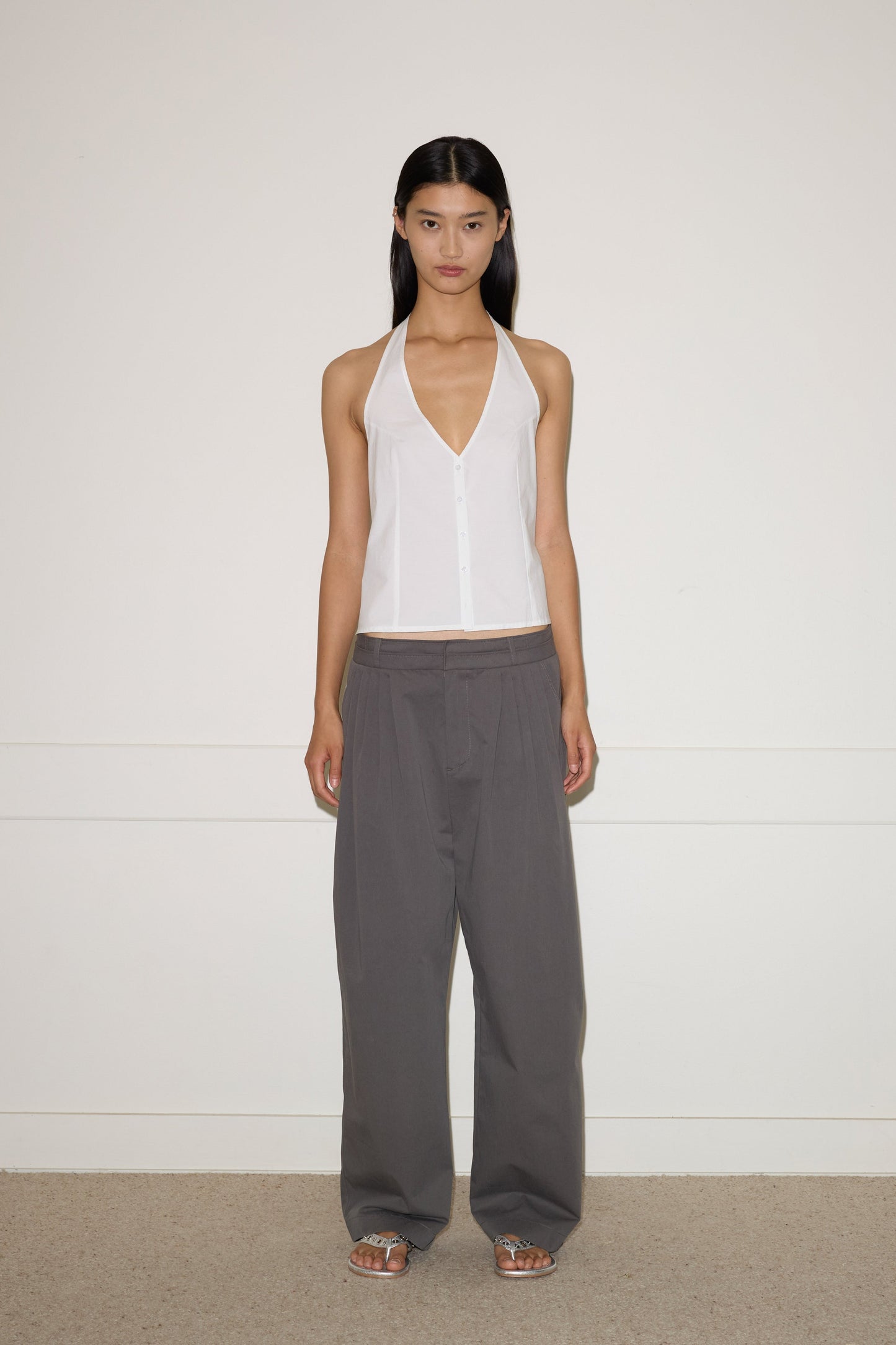 A woman with straight dark hair and a neutral expression stands against a plain wall, wearing a white halter-style sleeveless top, Deiji Studios Two Layer Pant - Charcoal in a loose fit, and silver sandals.