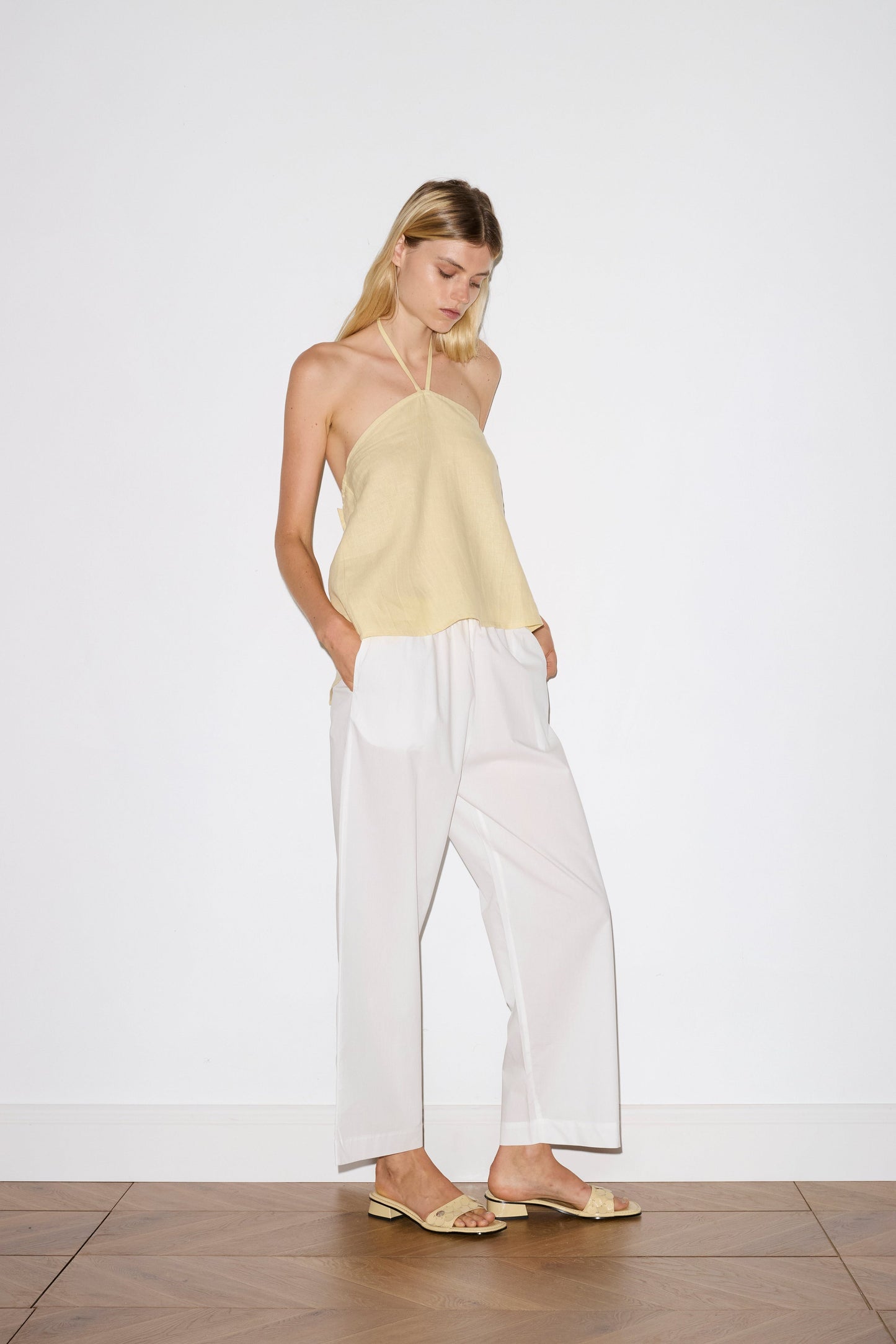 blonde haired female model wears a yellow relaxed halter top features flattering straight neckline, fine tie halter straps and snap closure at back with button detail, worn with a white ankle length pant features a soft minimal elastic waist with side seam pockets in a straight leg.