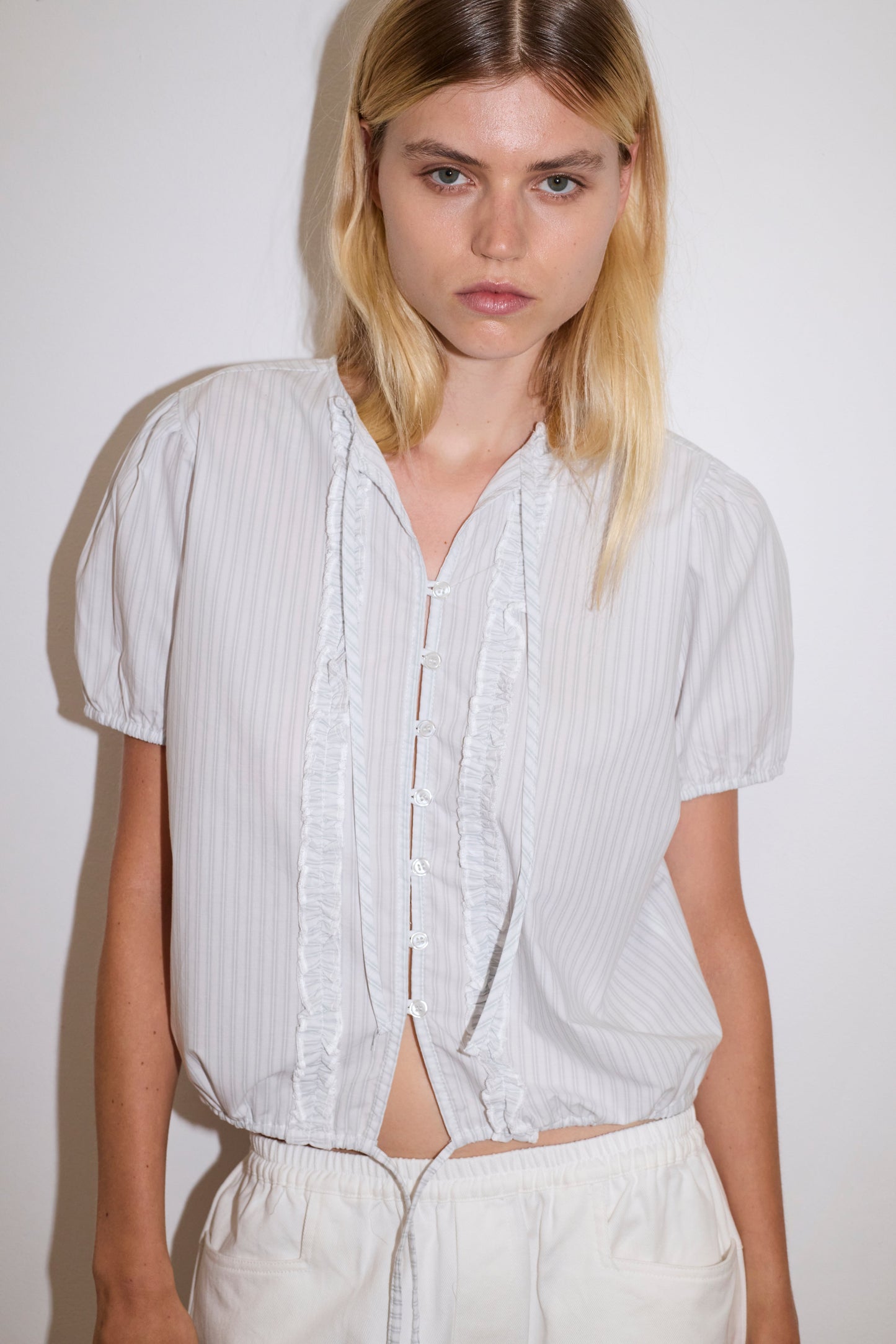 A woman with long blonde hair wears the Double Frill Top - Sage Stripe by Deiji Studios and white pants, standing against a plain white wall, looking directly at the camera with a neutral expression.