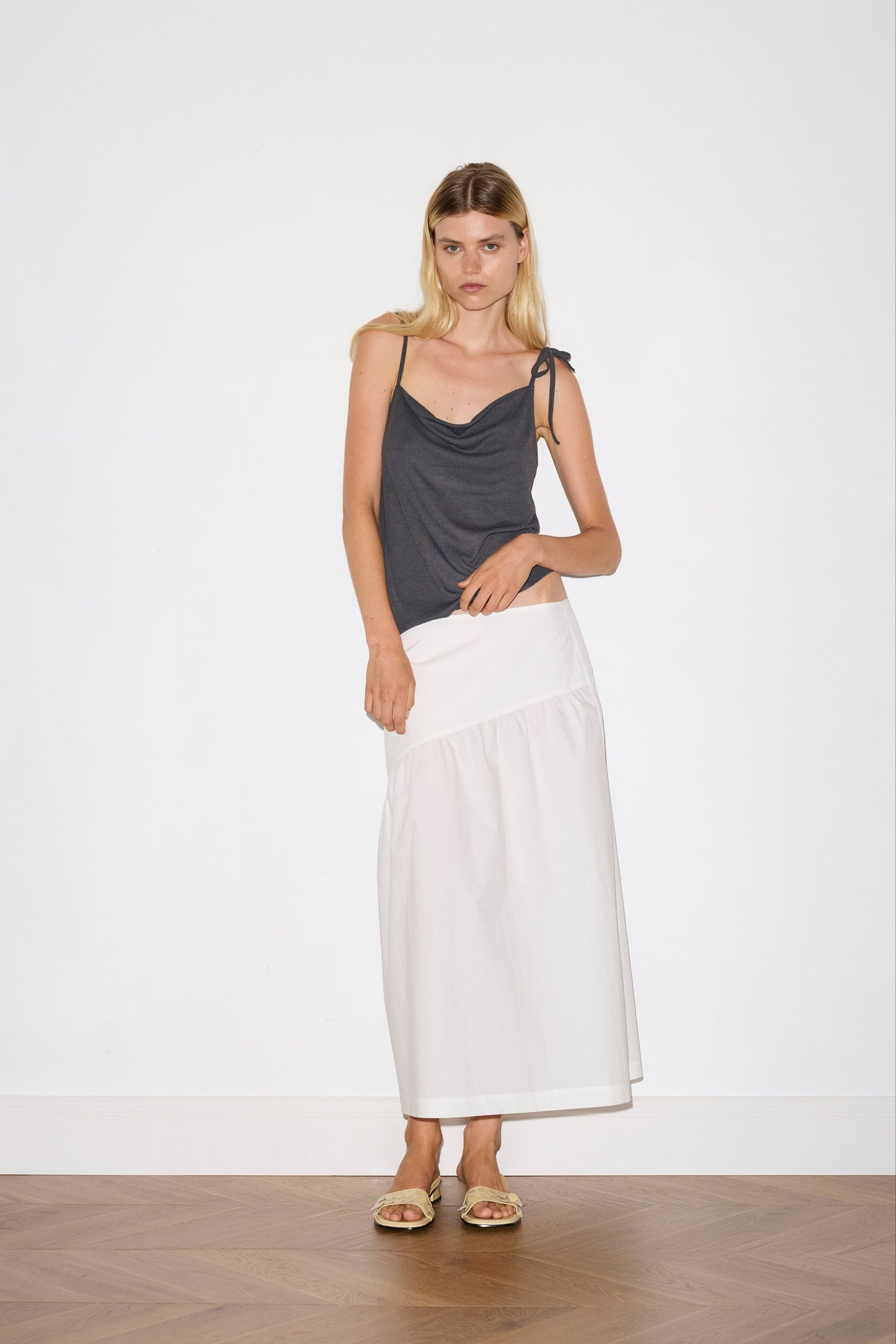 blonde female model wears a sheer fine knit relaxed tank with a fold over cowl neckline and low draping back, features self fabric rouleau straps, baby lock hem finishes and a asymmetric bottom detail, worn with a midi length white skirt
