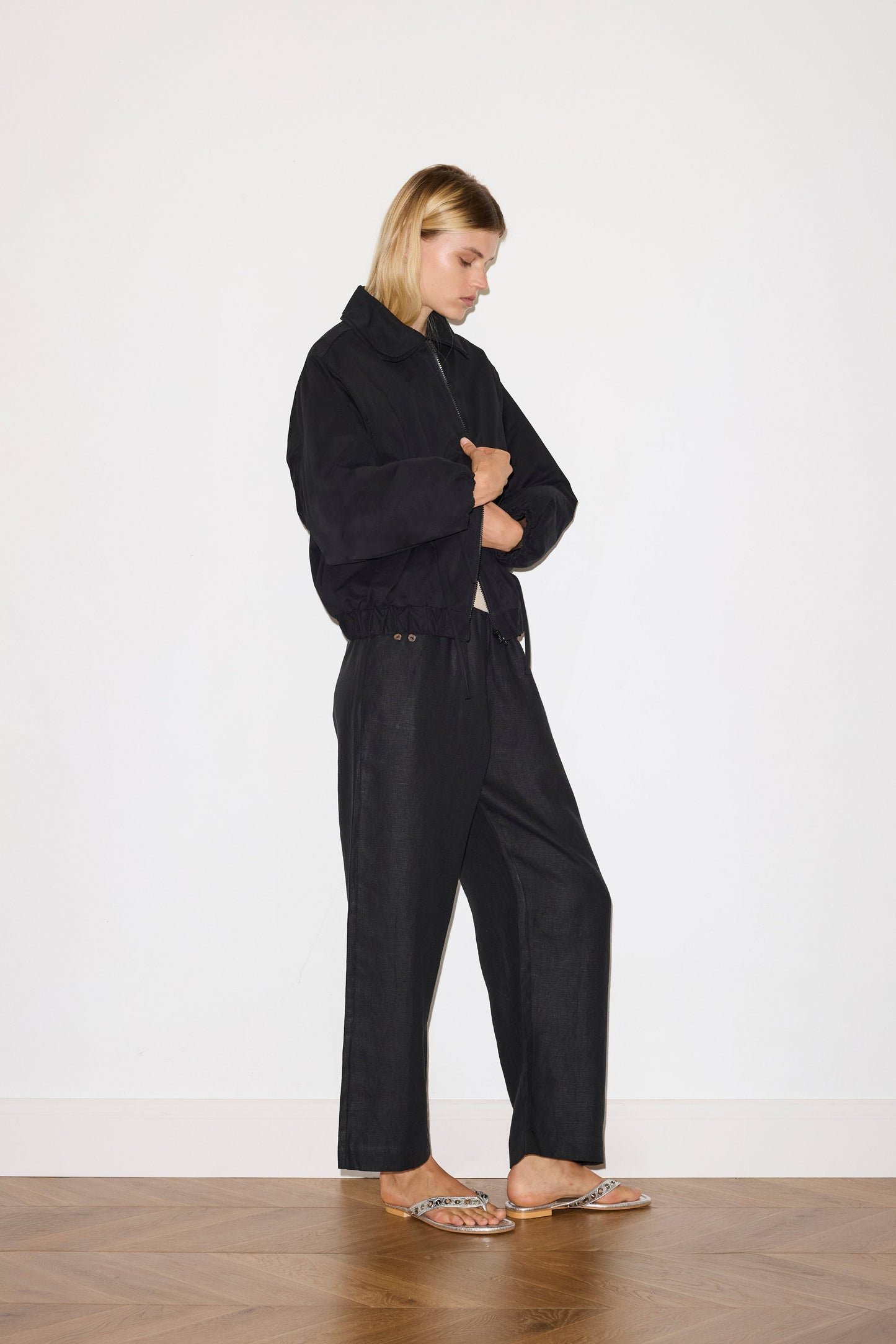 A person with short blonde hair stands indoors against a white wall, wearing the Double Layered Jacket - Black, matching black pants, and flat sandals. Arms crossed and eyes down, they stand on a wooden floor.