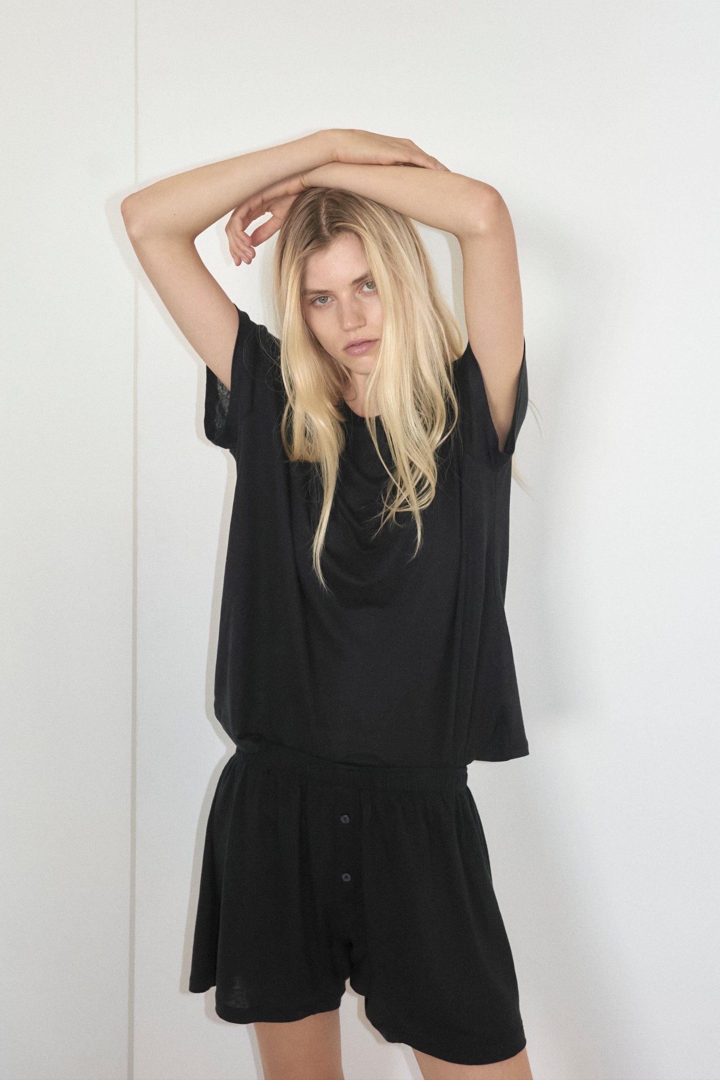A woman with long blonde hair stands against a white background, wearing Deiji Studios Soft Short in black paired with a matching black t-shirt, her hands are rested crossed over her head, she is looking directly at the camera with a neutral expression.