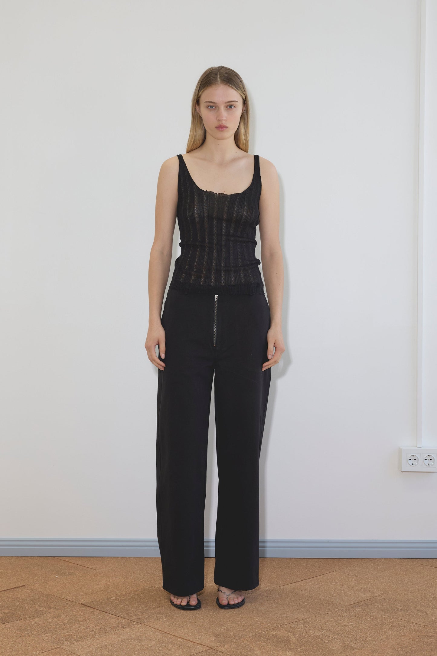 A woman stands indoors against a white wall, wearing the Open Work Tank - Black from Deiji Studios, high-waisted black pants with a front zipper, black sandals, and her straight hair loose.