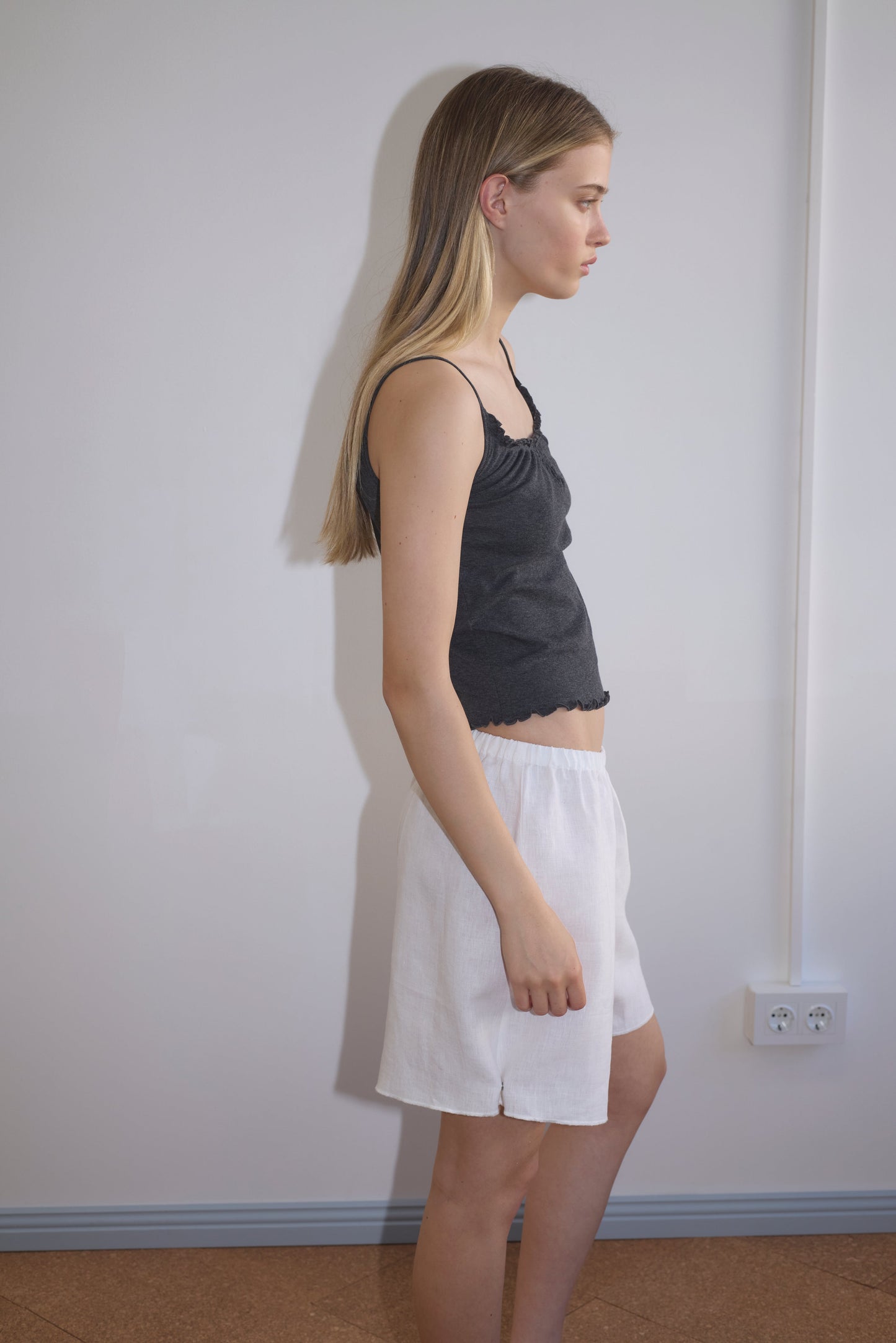 A young woman with long blonde hair stands in profile against a plain white wall, wearing a black tank top and the Press Stud Short - White from Deiji Studios, featuring an elastic waistline. She has a neutral expression and her arms are at her sides.