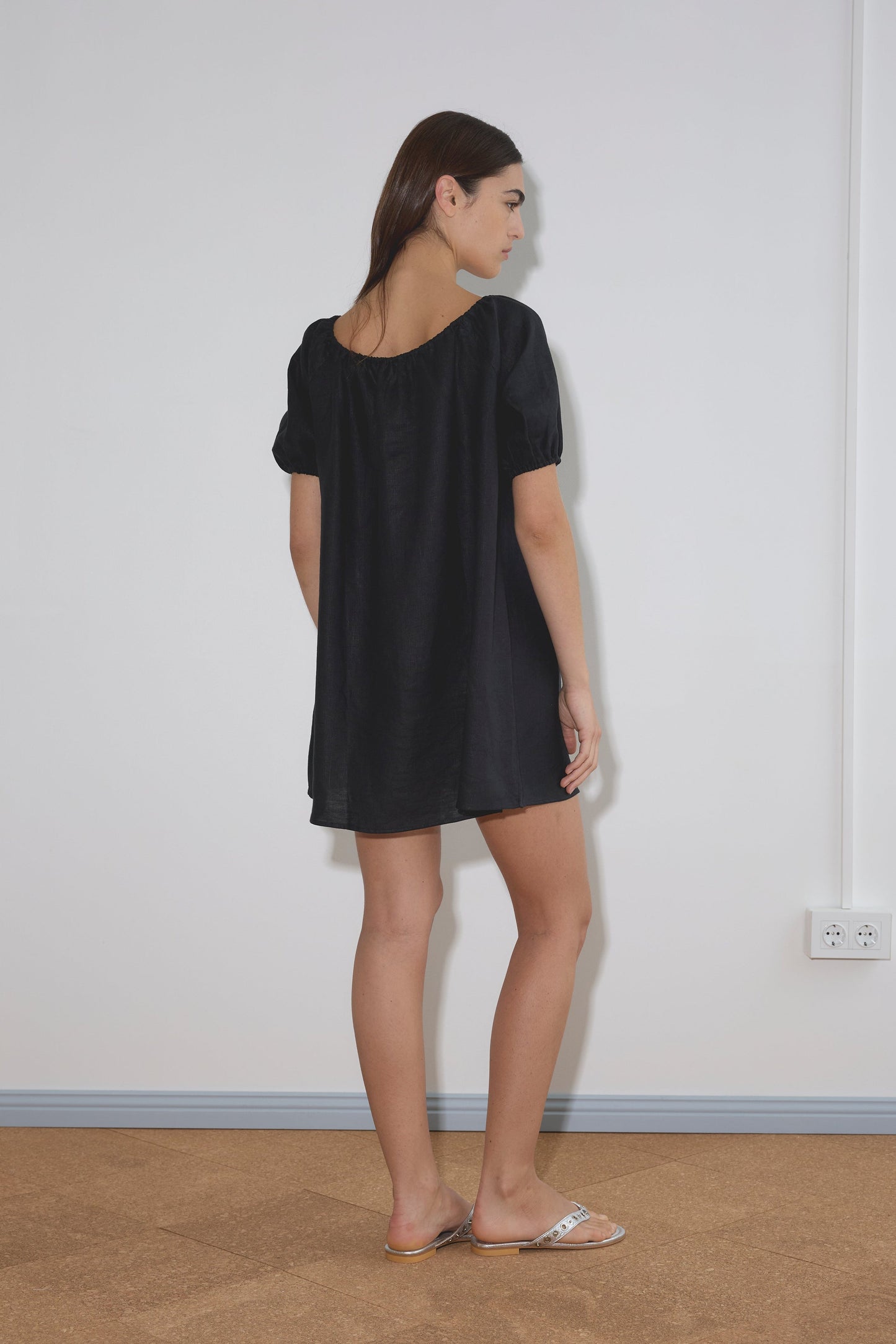 A woman stands indoors facing sideways, wearing Deiji Studios' Smock Dress - Black with short sleeves and flat sandals. She is in front of a plain white wall with a visible power outlet.