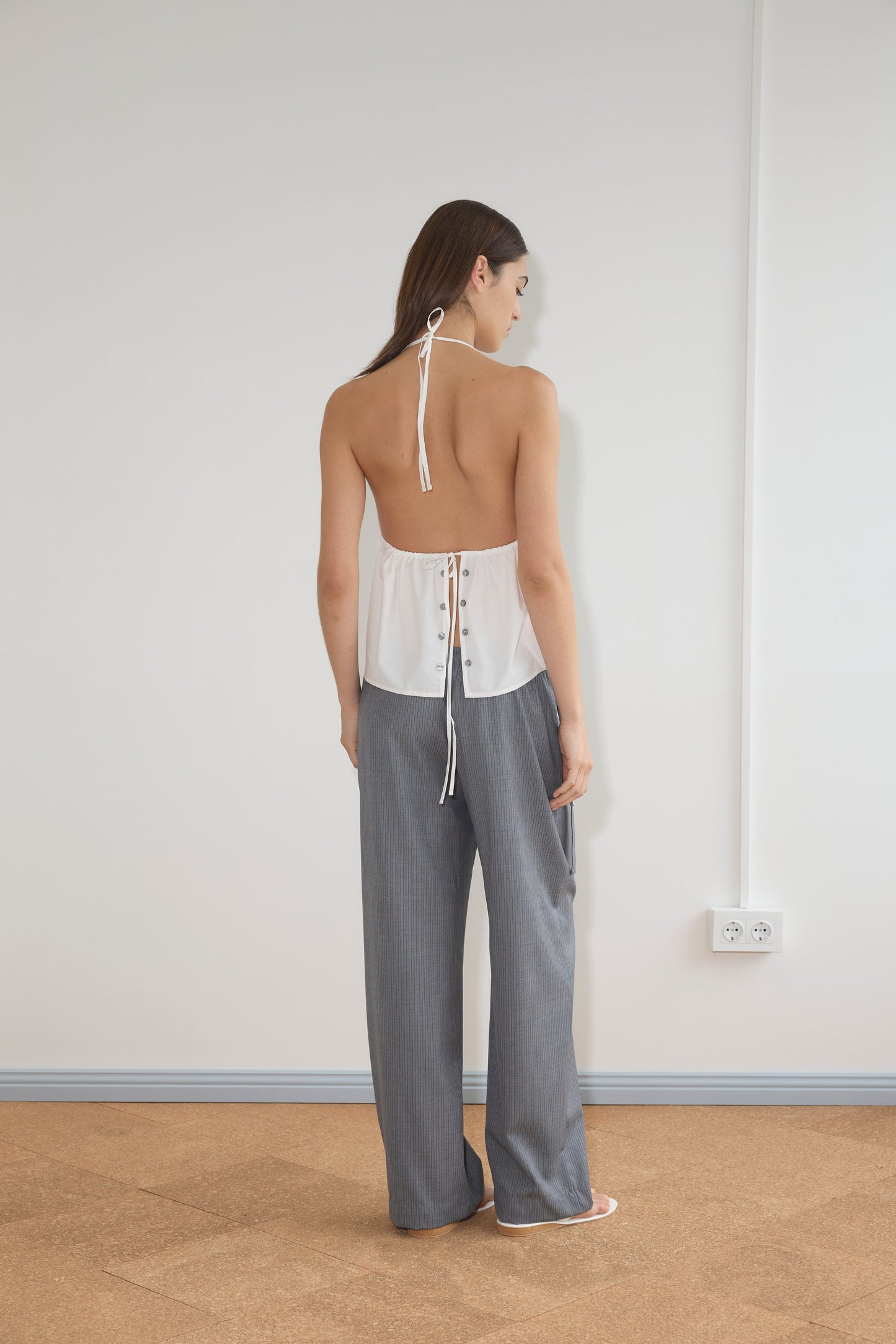 A woman stands facing a white wall, wearing the Button Halter - White with an open back and tie detail, paired with loose gray pants and white sandals. Her straight hair flows down as she faces away from the camera.