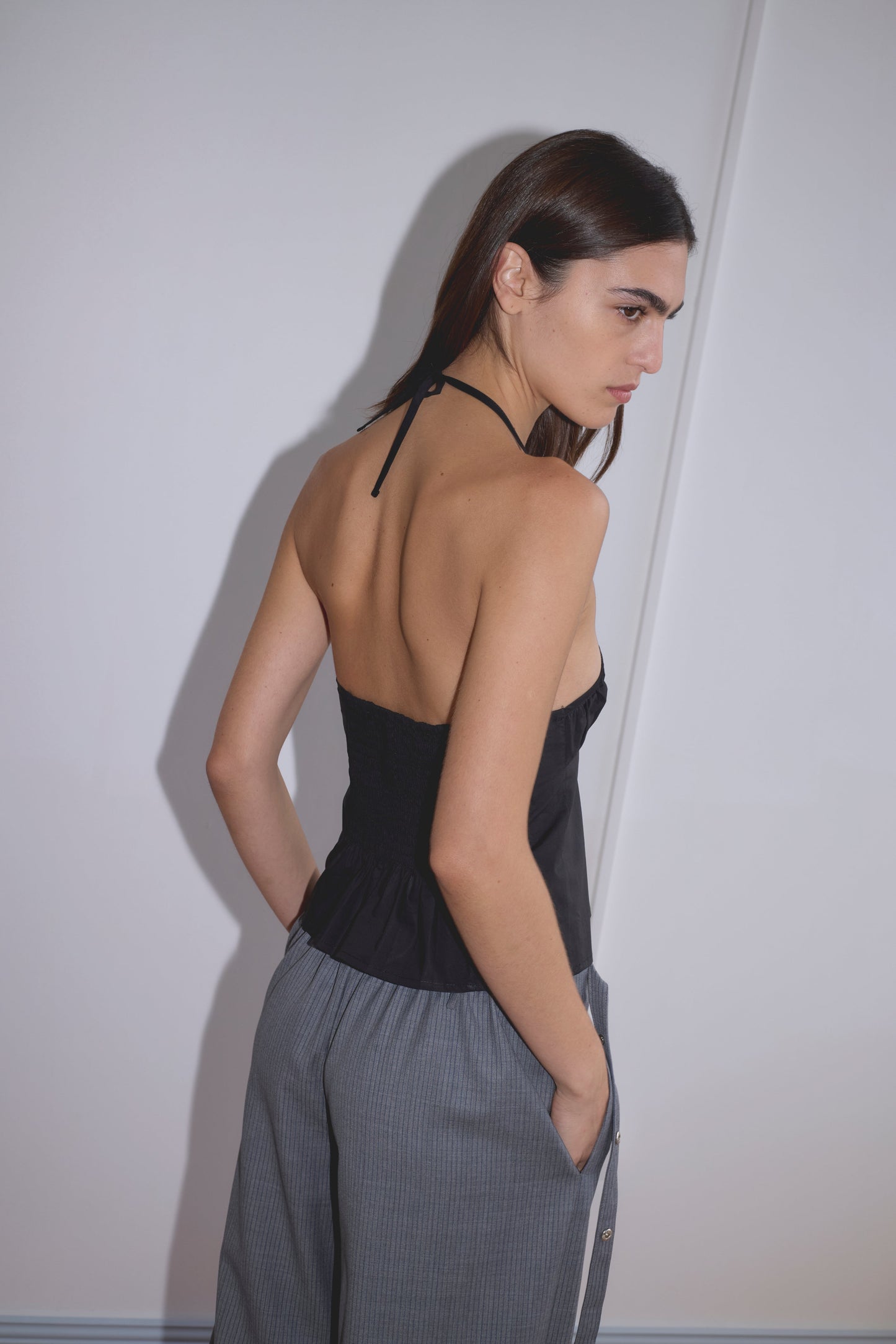 A woman with long brown hair stands sideways, looking over her shoulder. She wears the Strap Top - Black by Deiji Studios, featuring an open back, paired with gray high-waisted pants, her hands in the pockets against a plain white wall.