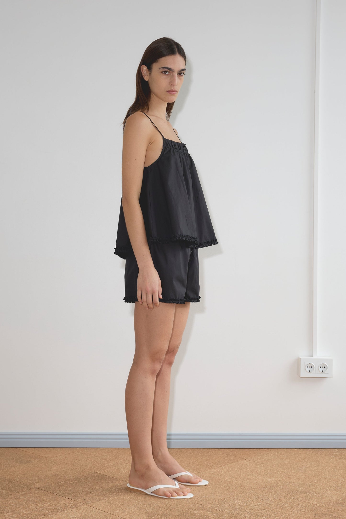 A woman stands sideways in a minimal room, wearing Deiji Studios' Yoke Set - Black—loose sleeveless top and matching shorts—paired with white slide sandals. The setting features a white wall, power outlet, and cork flooring.