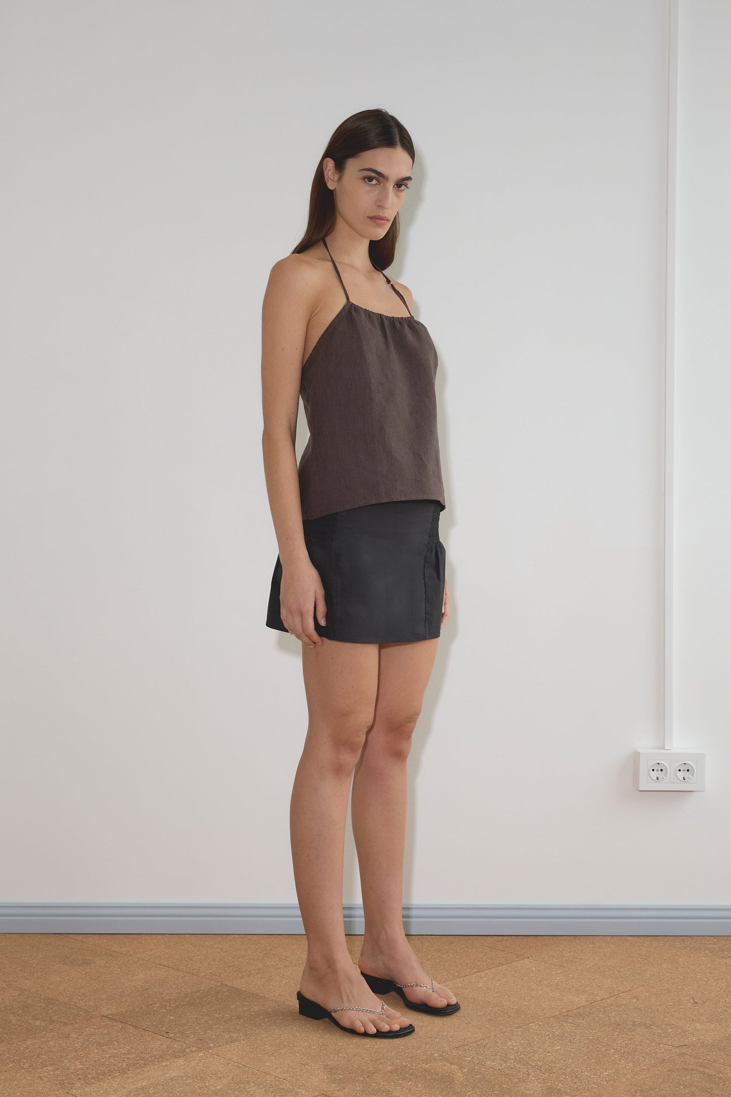 A woman stands indoors against a white wall, wearing a brown halter top, the Shirring Skirt - Black with subtle shirring details from Deiji Studios, and black sandals. She has straight dark hair and a neutral expression.