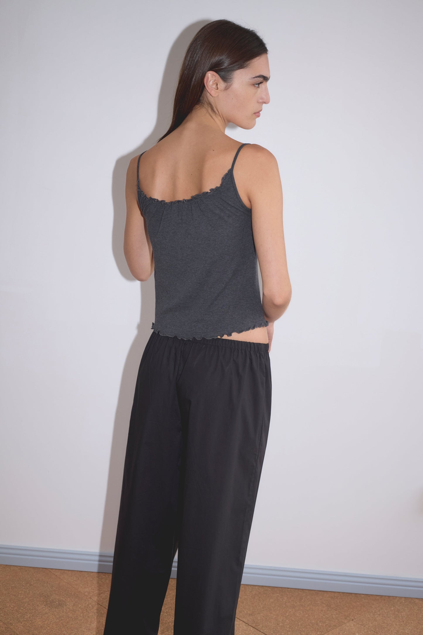 A woman with straight brown hair faces away from the camera, wearing a dark gray sleeveless Deiji Studios top and loose Flounce Pant - Black, against a plain white wall and light brown floor.