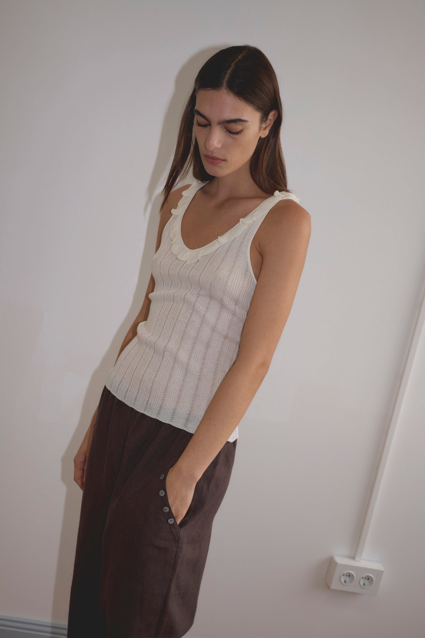 Model wears the Neck Ruffle Knit - Off White by Deiji Studios, featuring a delicate neck ruffle, styled with a dark brown midi skirt with side buttons, standing against a white wall.