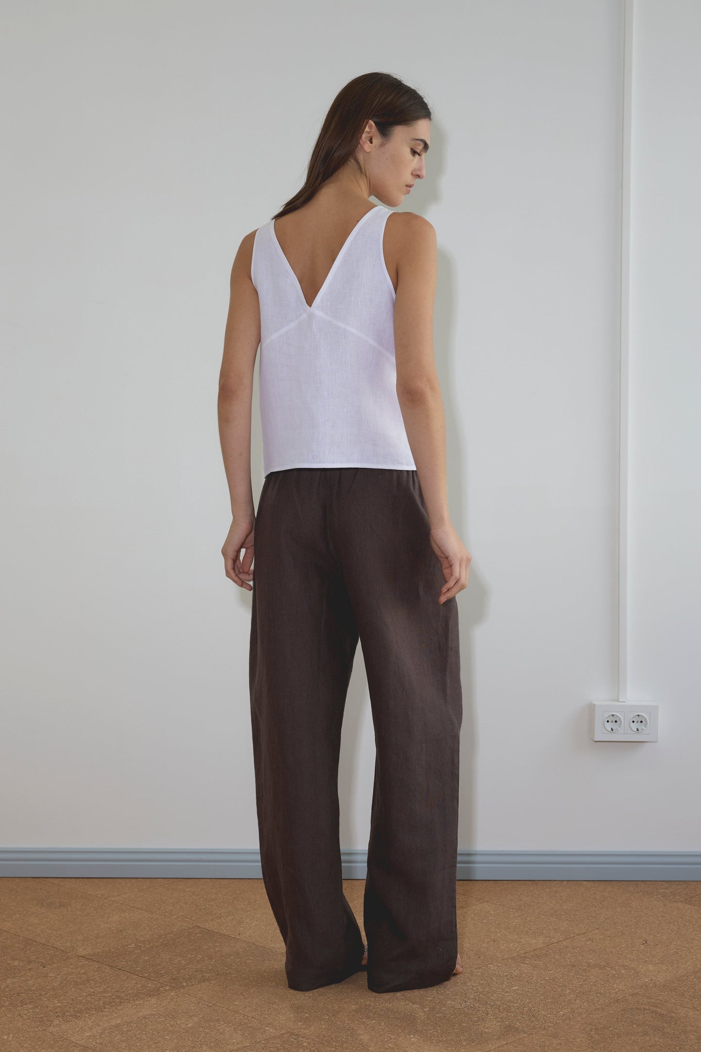 A woman stands indoors facing away, wearing the Deiji Studios Two Part Top - White with a deep v-neckline and loose dark brown pants, on a light brown floor near a white wall with two electrical outlets.
