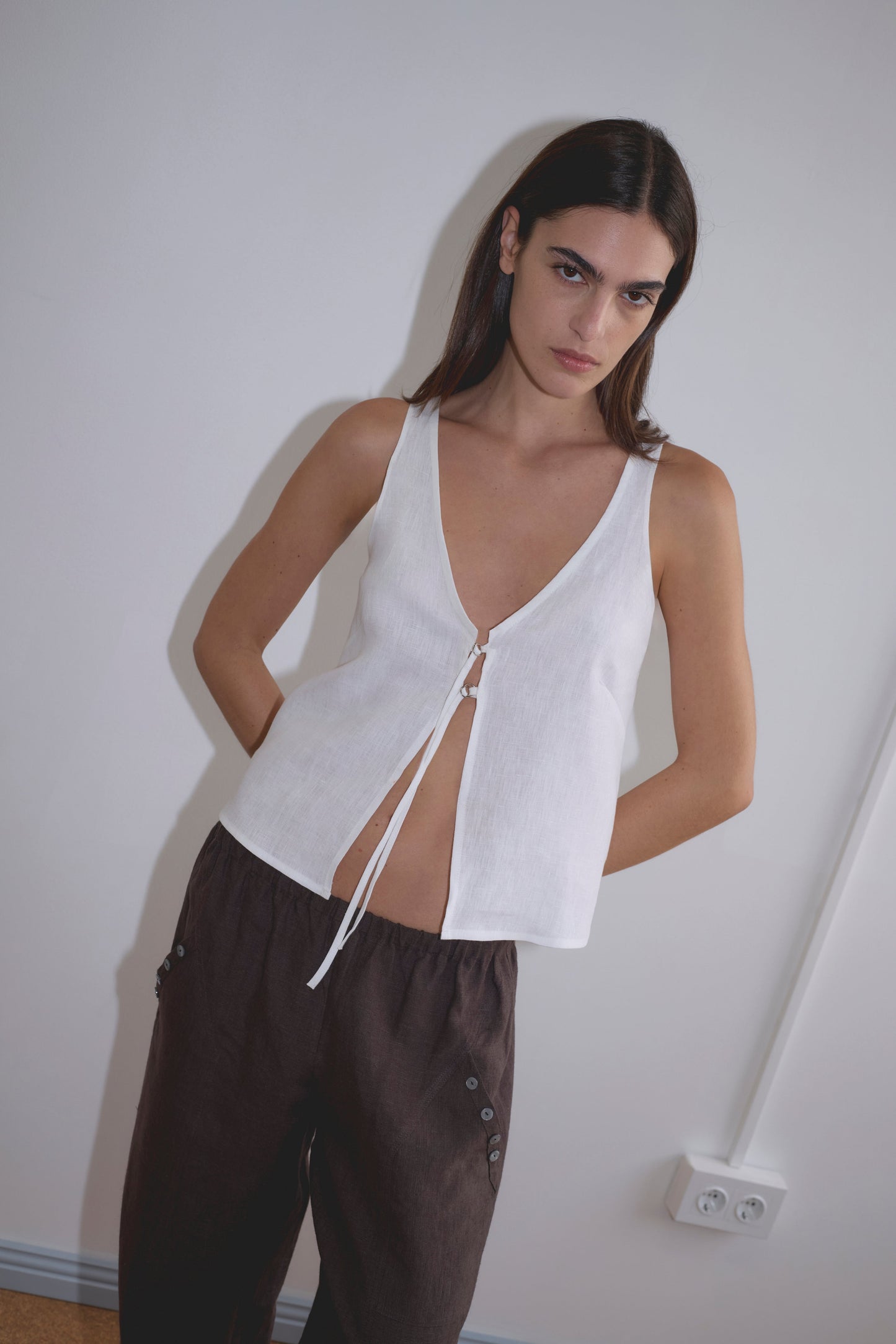A woman with long dark hair leans against a white wall, wearing the Deiji Studios Two Part Top in white, sleeveless and tied at the front, paired with loose dark brown button-detail pants. An electrical outlet is visible near the floor.