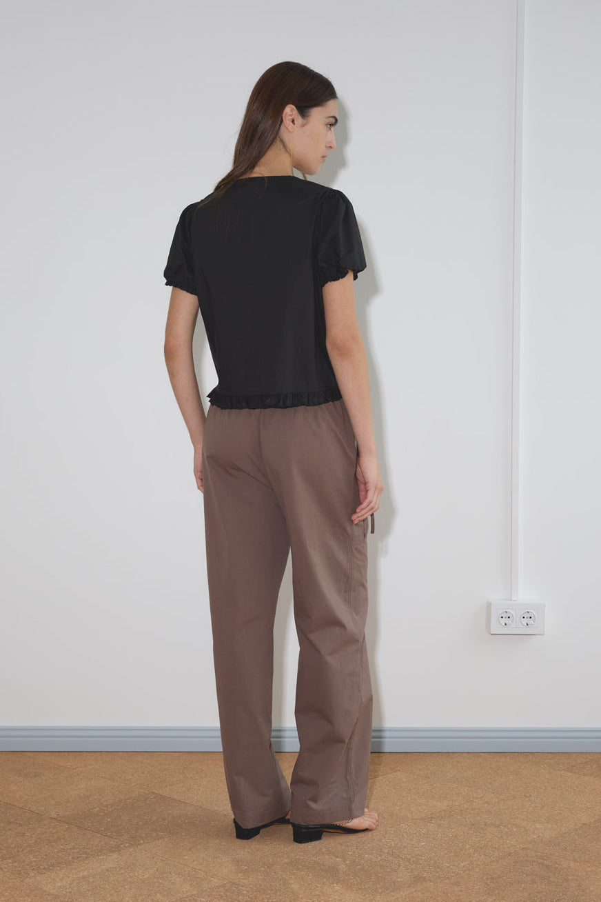 A woman stands indoors with her back to the camera, wearing the Pintuck Top - Black by Deiji Studios and loose brown pants. The room has plain white walls, a visible electrical outlet, and a beige floor.