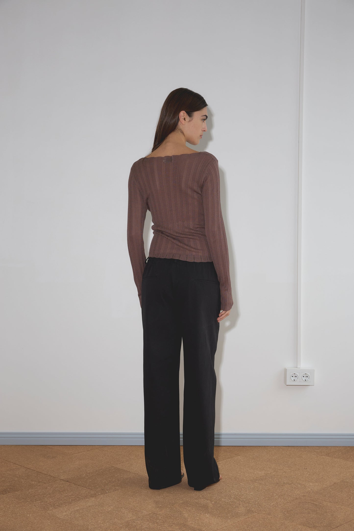 A woman with long brown hair stands facing a white wall, wearing a brown long-sleeve top and the Zip Pant - Black by Deiji Studios. A power outlet is visible on the wall near her right side.