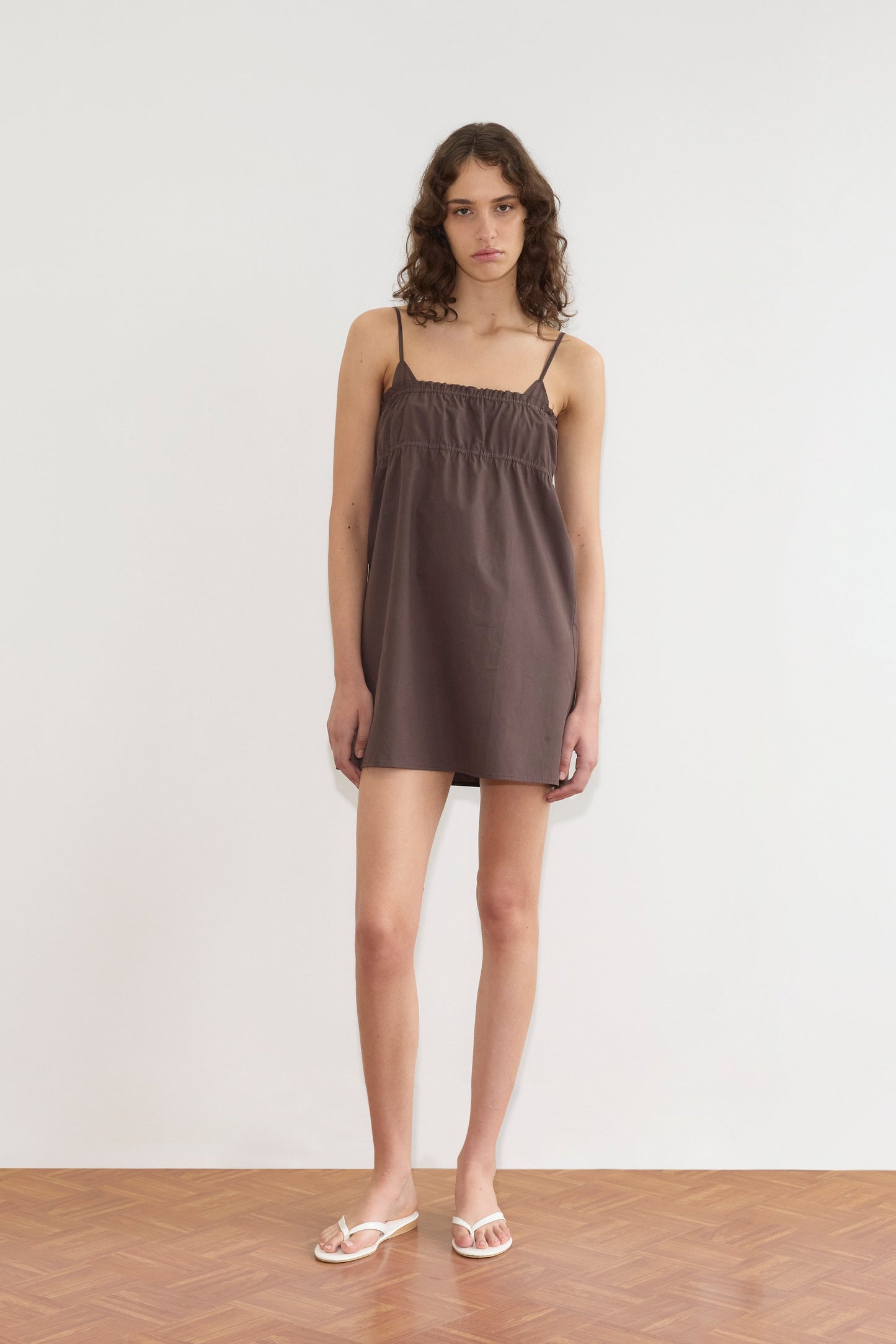 A person with curly brown hair wears the Edge Dress - Sepia from Deiji Studios and white sandals, standing against a plain white wall on a wooden floor.