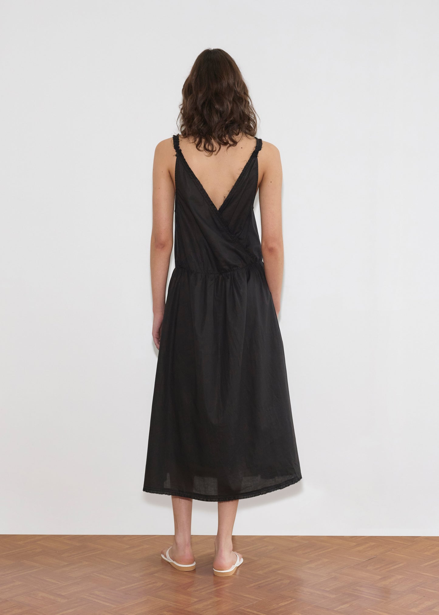 A woman with shoulder-length wavy hair stands facing a white wall, wearing the Crossed Dress - Black by Deiji Studios with a crossover v-neckline and V-back, paired with light-colored flat sandals on a wooden floor.