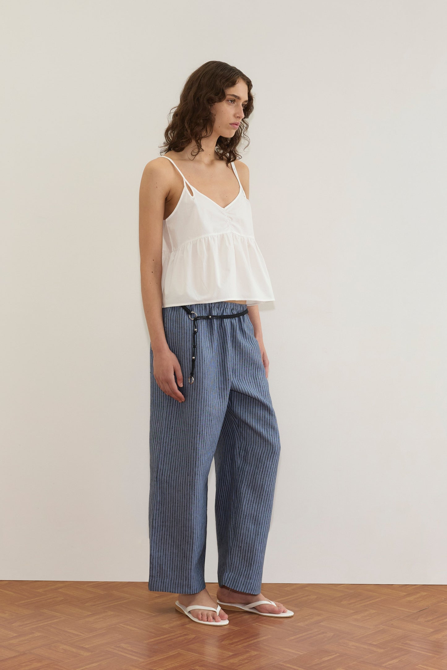 A person with wavy brown hair wears the Bind Top - White by Deiji Studios, loose blue striped pants, and white sandals, standing on a parquet floor against a plain white wall.