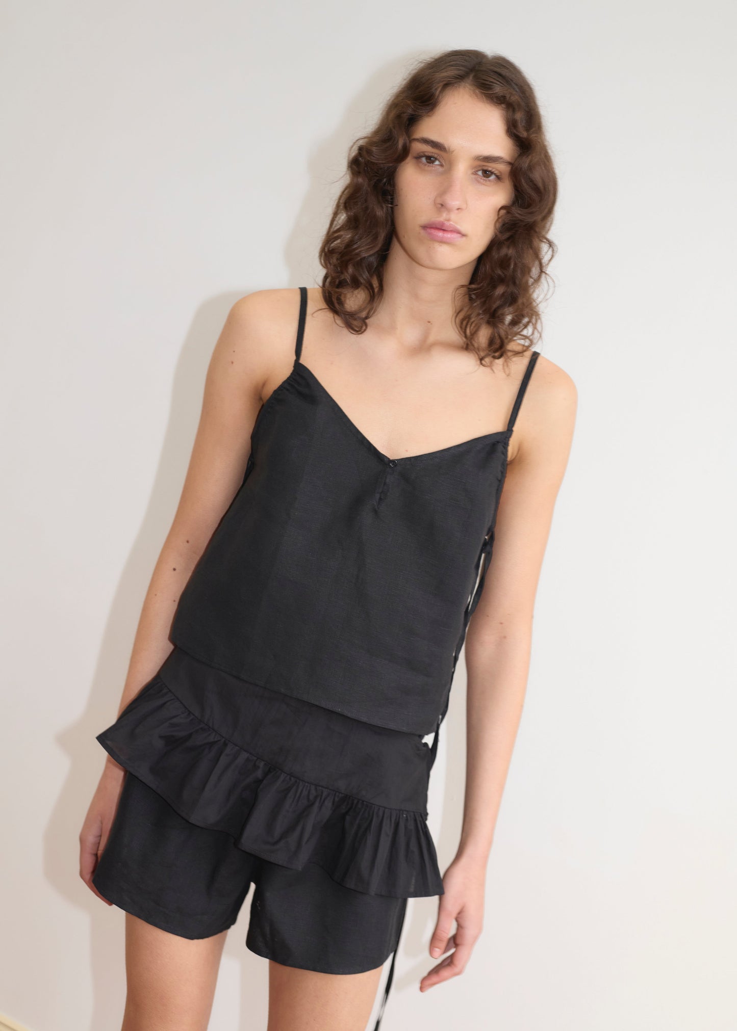 A person with wavy brown hair models the Flounce Belt - Black, styled with a sleeveless black top and matching shorts, against a plain white background.