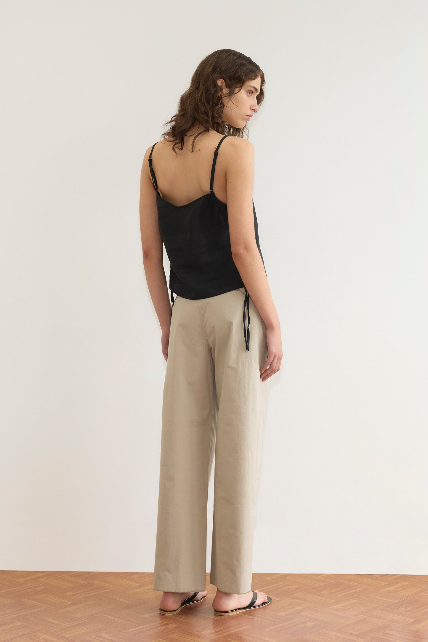A woman with wavy brown hair stands on a wooden floor, facing away, wearing a V Neck Cami - Black, beige wide-leg pants, and black flip-flops against a plain white background.