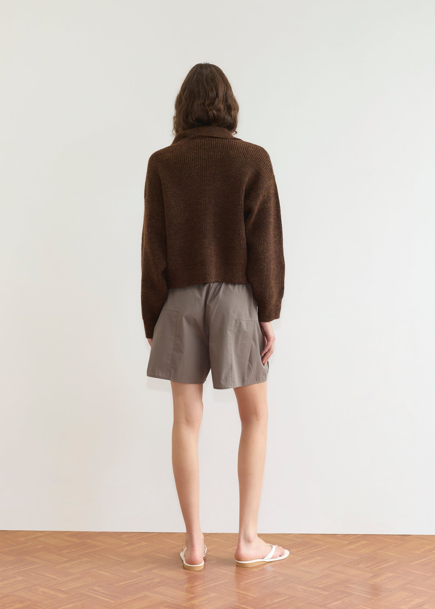 A person with wavy brown hair stands facing away, wearing a Deiji Studios Double Layer Cardigan in brown, loose gray shorts, and white flip-flops against a plain white wall and wooden floor.