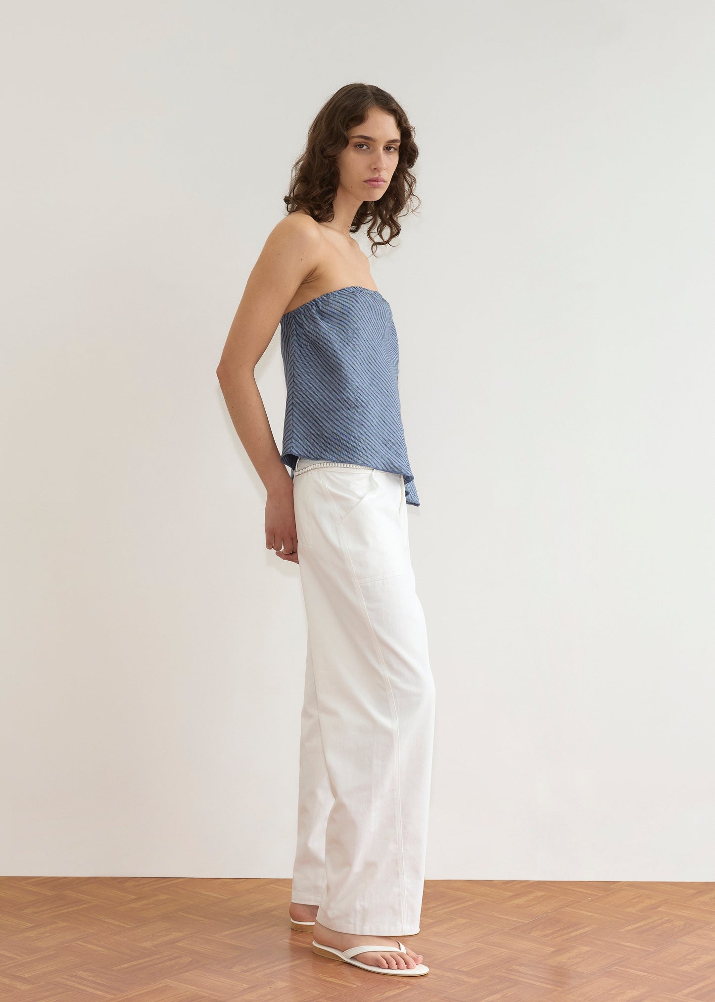 A person with wavy brown hair stands indoors against a white wall, wearing a blue strapless top, white wide-leg pants styled with the Ring Loop Belt - White, and white sandals on a wooden floor.