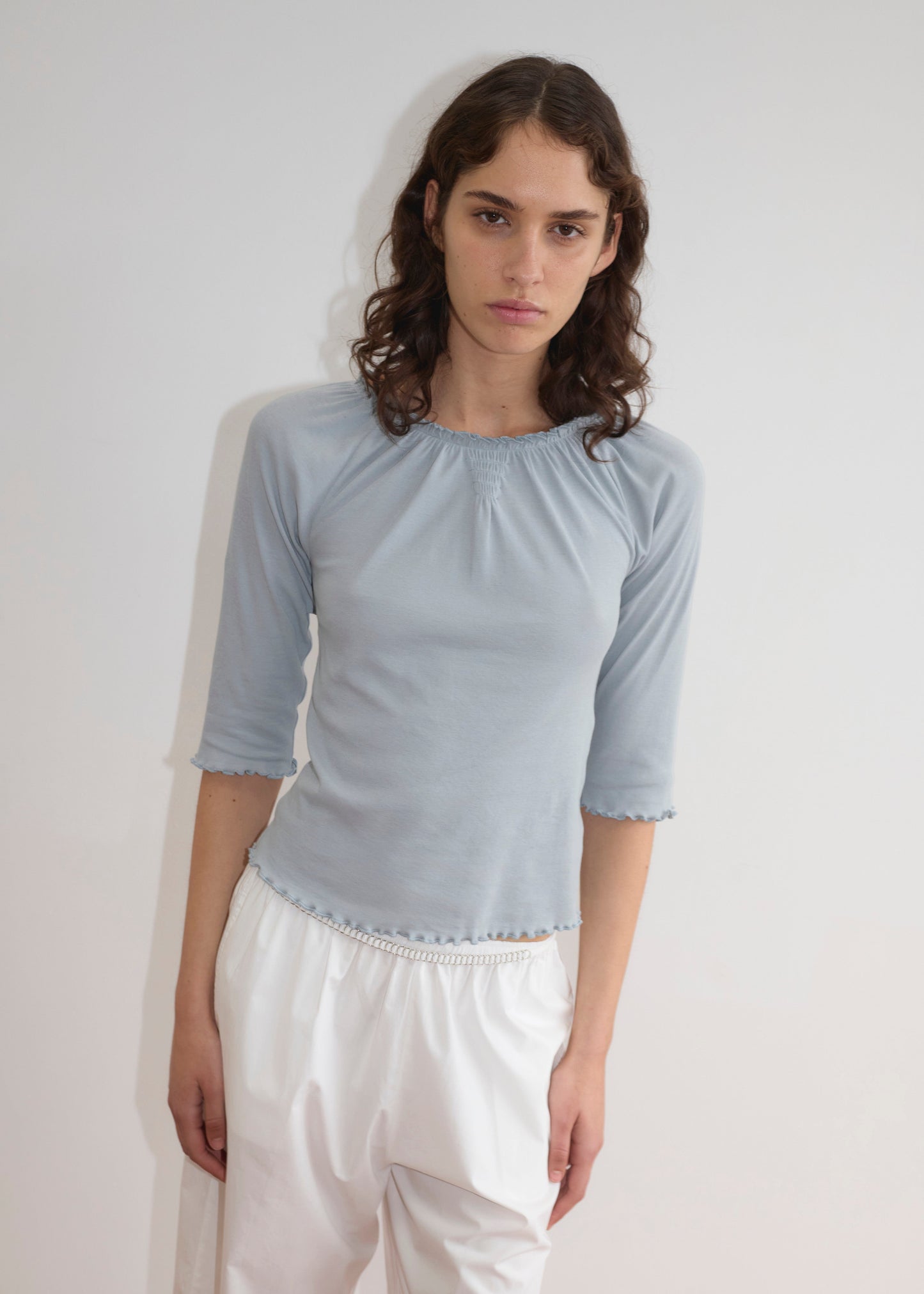 A person with wavy brown hair wears a Deiji Studios Cotton Flounce 3/4 Sleeve top in baby blue and loose white pants, standing against a plain white background with a neutral expression.
