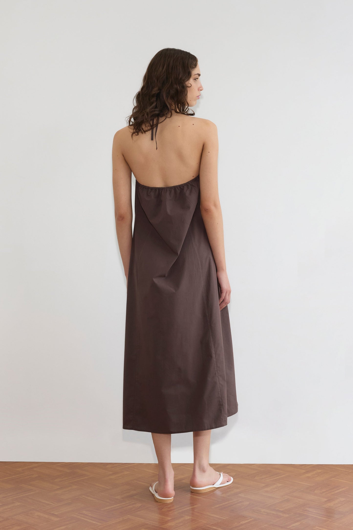 A woman with wavy brown hair, wearing the Seamed Halter Dress - Sepia and white sandals, stands on a wooden floor facing away from the camera against a plain white wall.