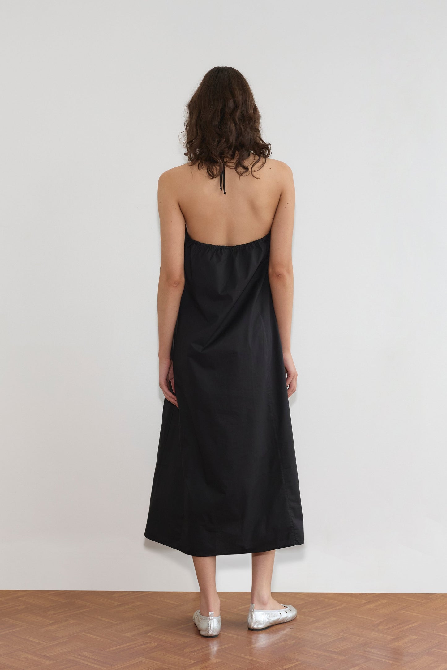 A woman with wavy brown hair stands facing a white wall, wearing the black Seamed Halter Dress and silver flats on a wooden floor.