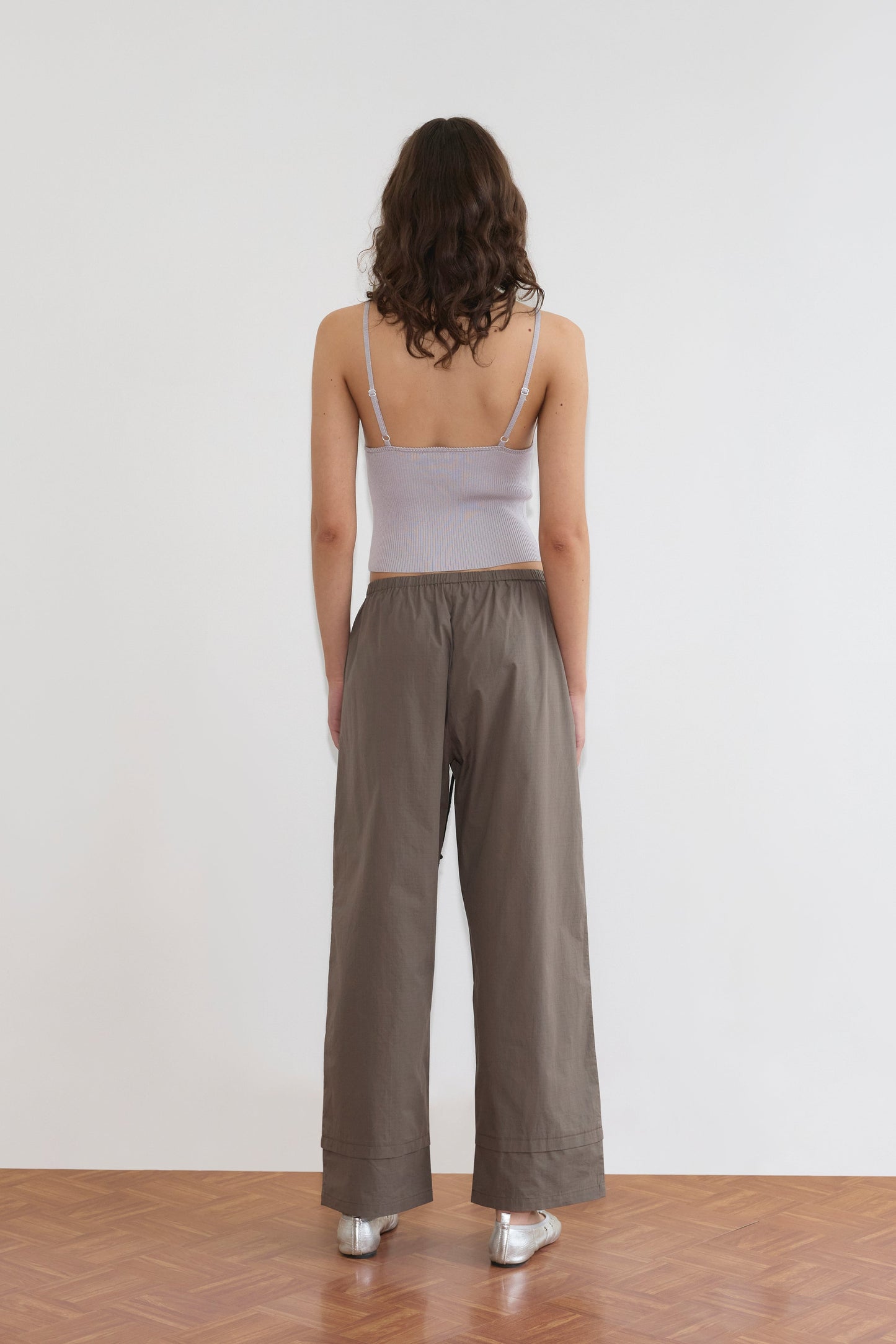 A woman with wavy brown hair faces away, wearing the Cross Panel Knit Top - Soft Lilac by Deiji Studios, paired with loose dark gray pants and silver shoes, standing on a wooden floor against a plain white wall.