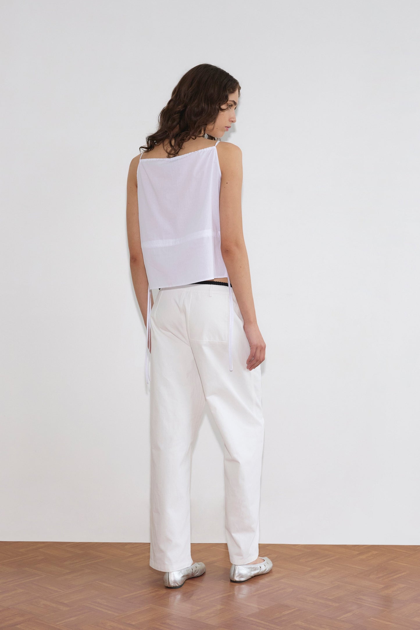 A person with wavy brown hair faces away, wearing a sleeveless white top with side ties, Pocket Jean - White, and silver shoes, set against a plain white wall and wooden floor.